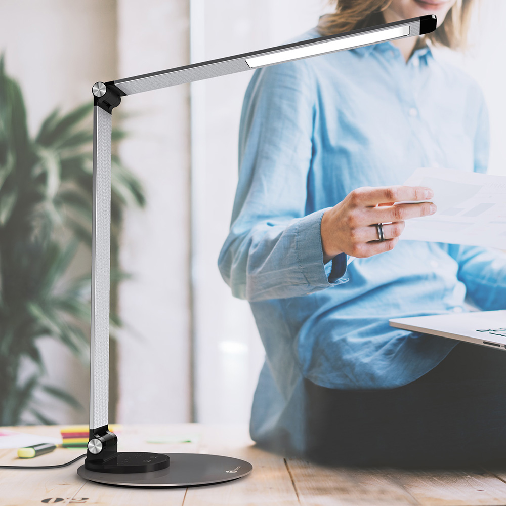 Eine Schreibtischlampe mit silbernem Arm und rundem Sockel steht auf einem Holztisch. Eine Person in blauem Hemd im Hintergrund.