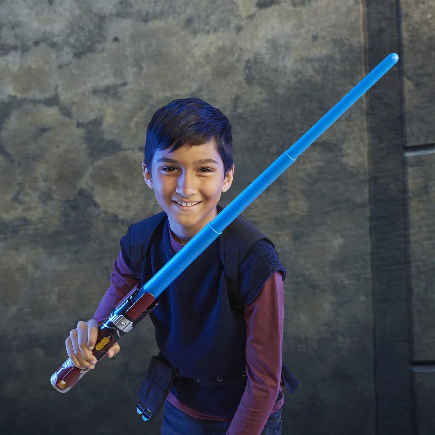 Un niño sonriente sostiene un sable de luz azul. Lleva un chaleco oscuro sobre una camisa granate. El fondo es una pared gris texturizada.