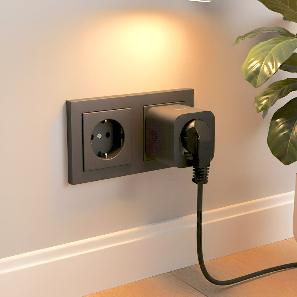 A dark gray wall outlet with a black plug inserted. A black cable runs down. A green plant is on the right.