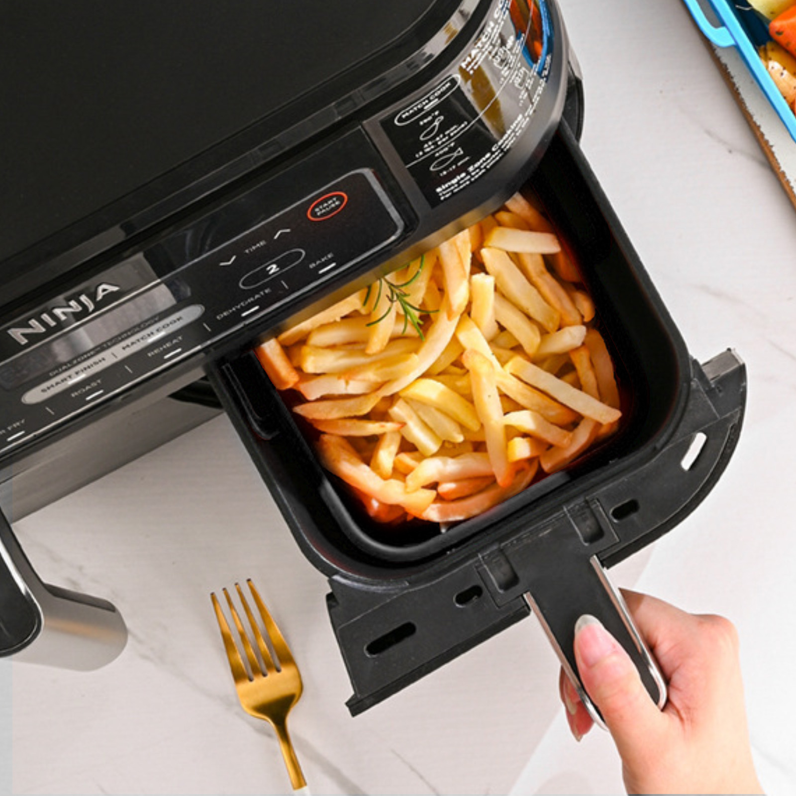 Ein Airfryer mit Pommes Frites. Eine Hand zieht die Schublade heraus. Eine goldene Gabel liegt auf der Theke.