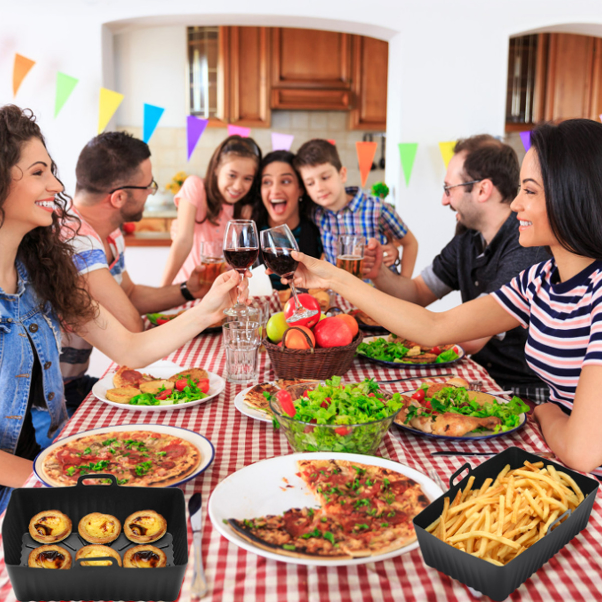 Eine Familie prostet mit Getränken an einem Tisch voller Essen, darunter Pizza, Salat, Pommes und Gebäck. Bunte Fahnen hängen oben.