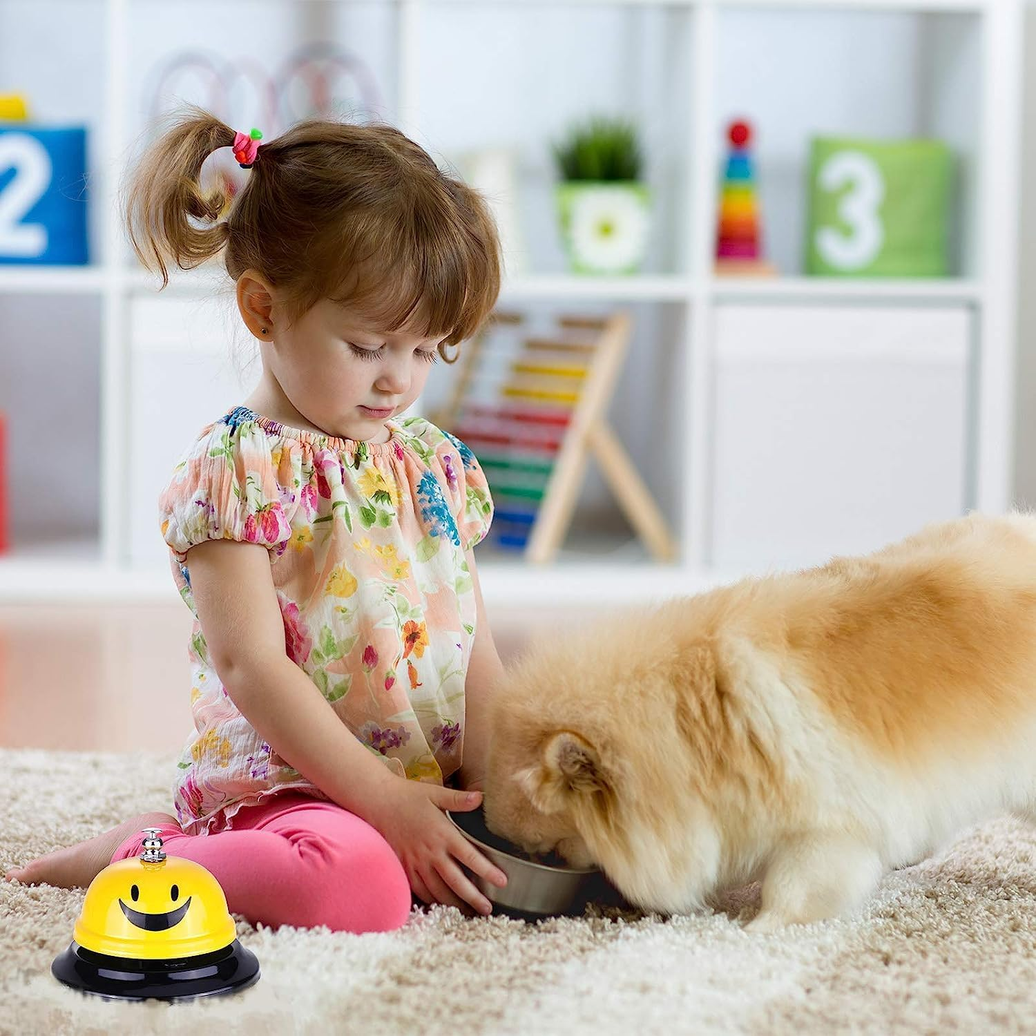 Ein Mädchen füttert einen flauschigen Hund. Auf dem Teppich steht eine gelbe Klingel mit Smiley-Gesicht. Im Hintergrund Regale mit Spielzeug.