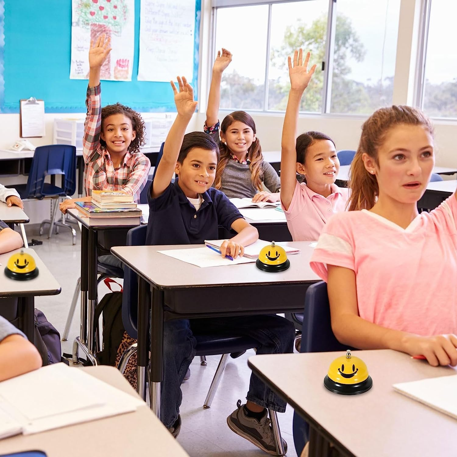 Schüler heben im Klassenzimmer die Hand. Schreibtische mit Smiley-Glocken. Blaue Tafel und Fenster im Hintergrund.