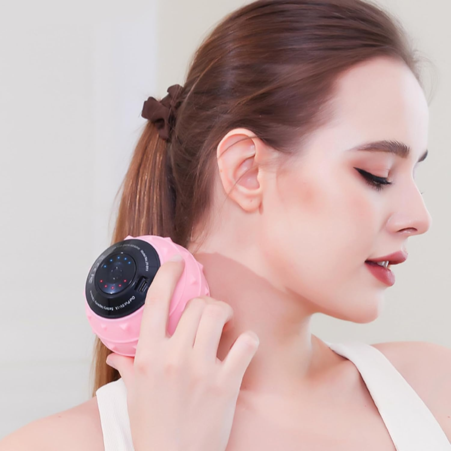 Eine Frau hält ein rosa Massagegerät an ihren Hals. Sie hat braune Haare, die zurückgebunden sind, trägt ein weißes Tanktop und roten Lippenstift.