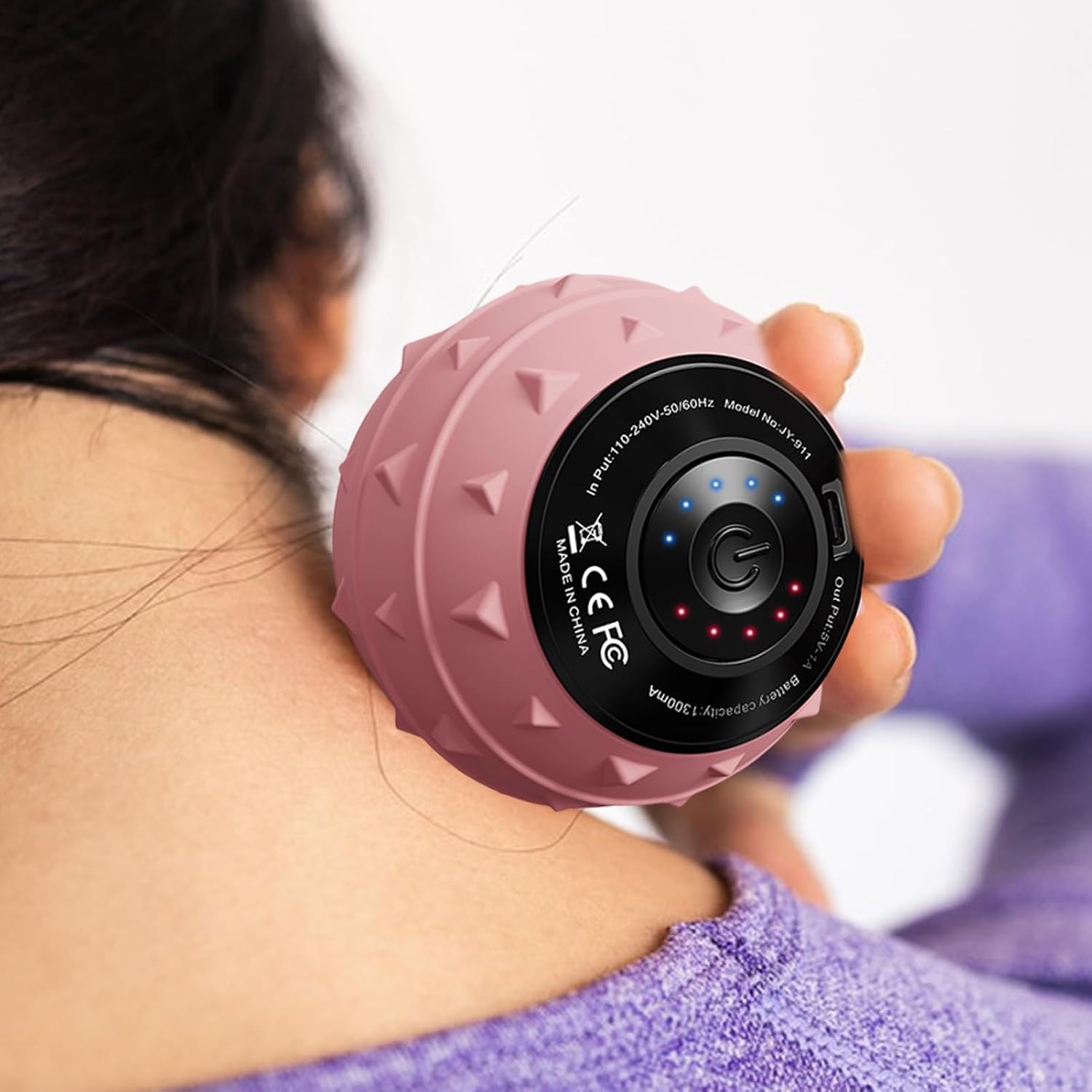 Eine Person hält einen rosa Massageball an ihrem Hals. Der Ball hat Stacheln und ein schwarzes Bedienfeld. Die Person trägt ein lila Hemd.