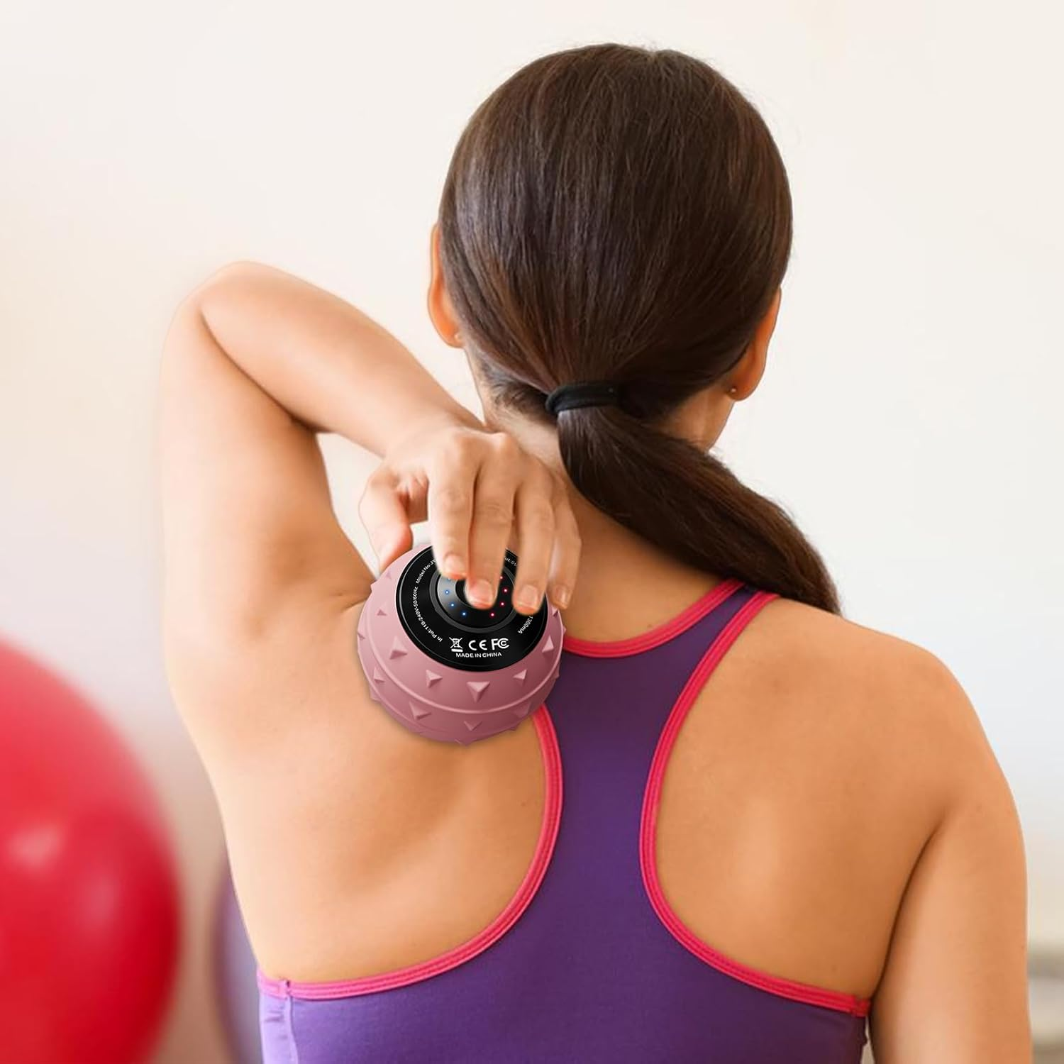 Eine Frau benutzt einen rosa Massageball auf ihrer Schulter. Sie trägt einen lila Sport-BH. Roter Gymnastikball im Hintergrund.