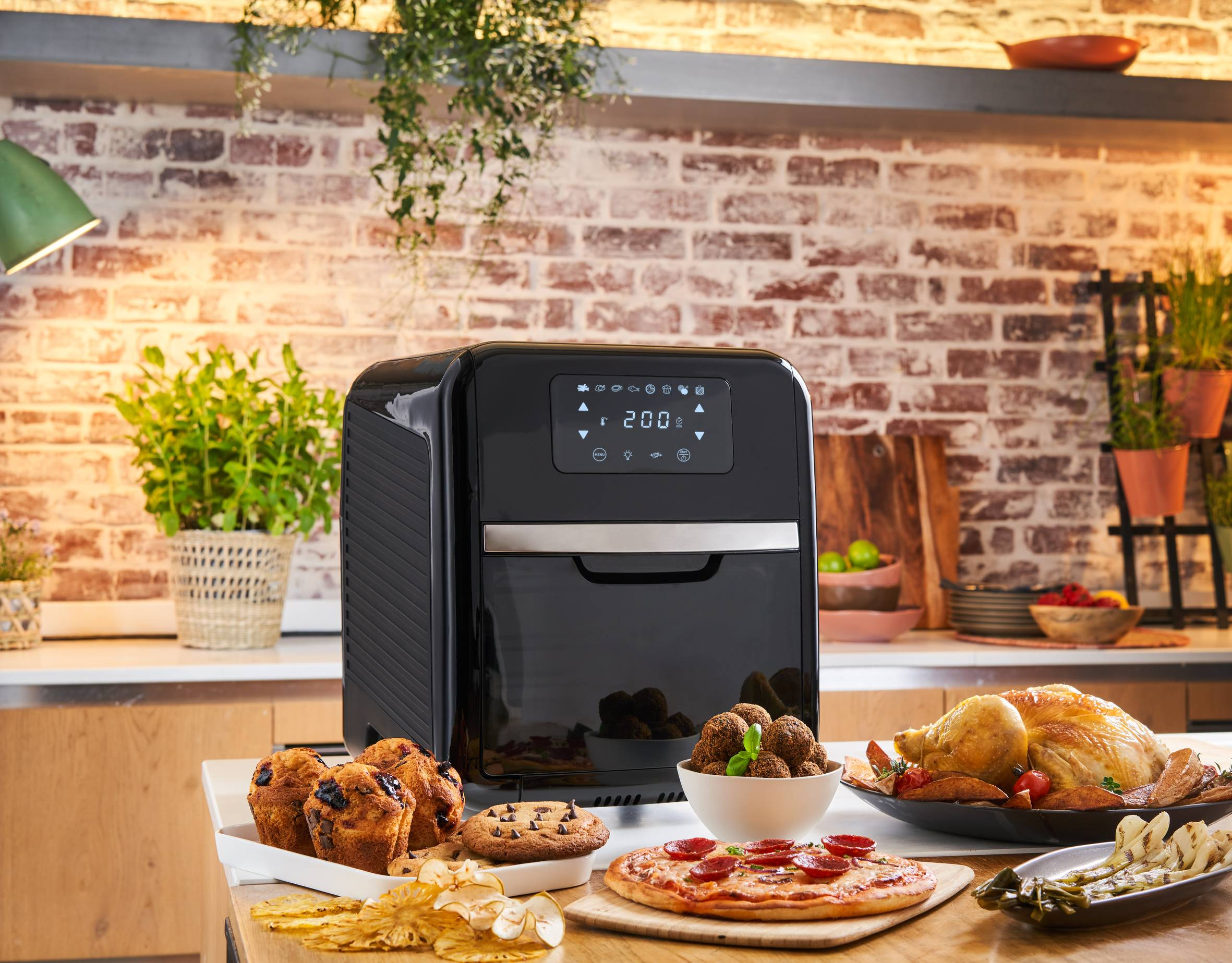 Zwarte airfryer, muffins, pizza, gebraden kip en ander voedsel op een houten tafel, voor een bakstenen muur.