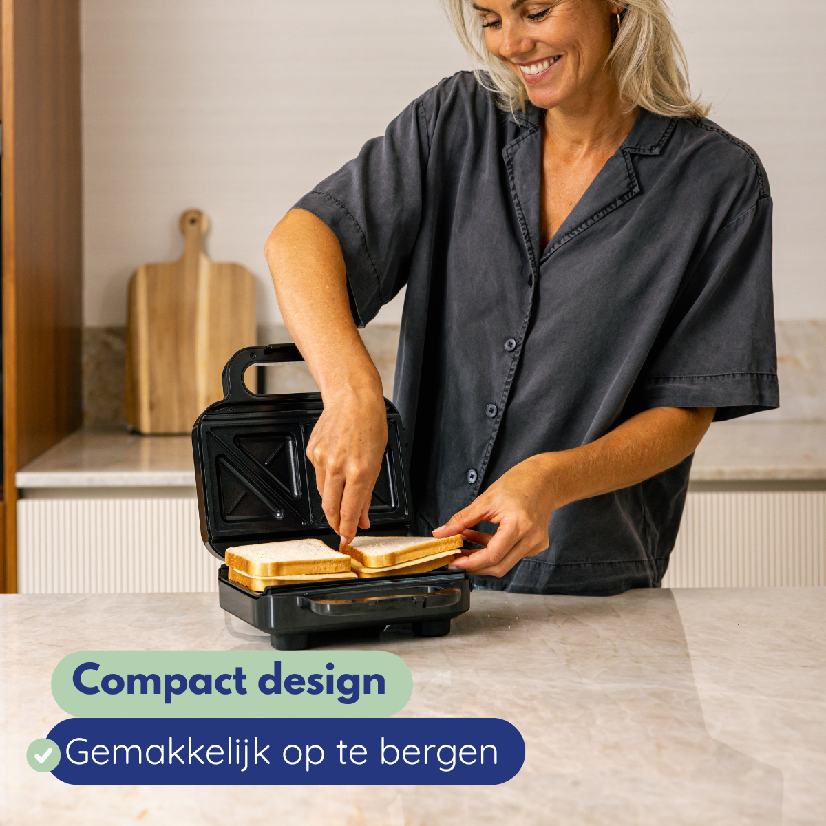 Een vrouw glimlacht terwijl ze brood in een zwarte sandwichmaker doet. De keuken heeft een houten snijplank op de achtergrond.