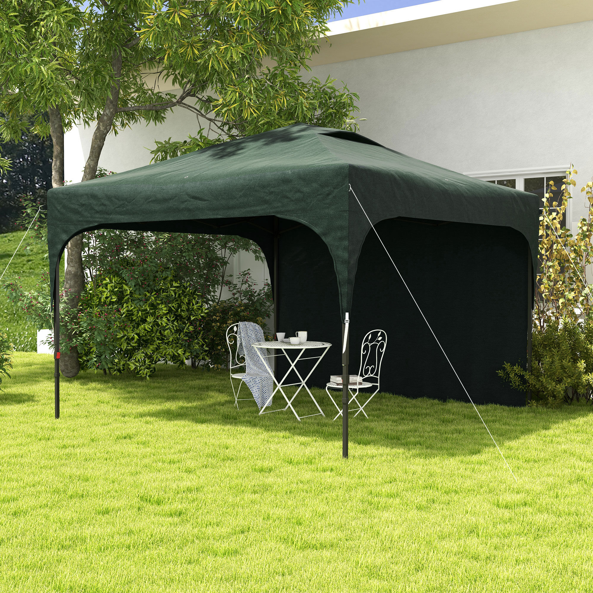 Toldo verde sobre mesa y sillas blancas sobre césped. Árboles y una casa al fondo.