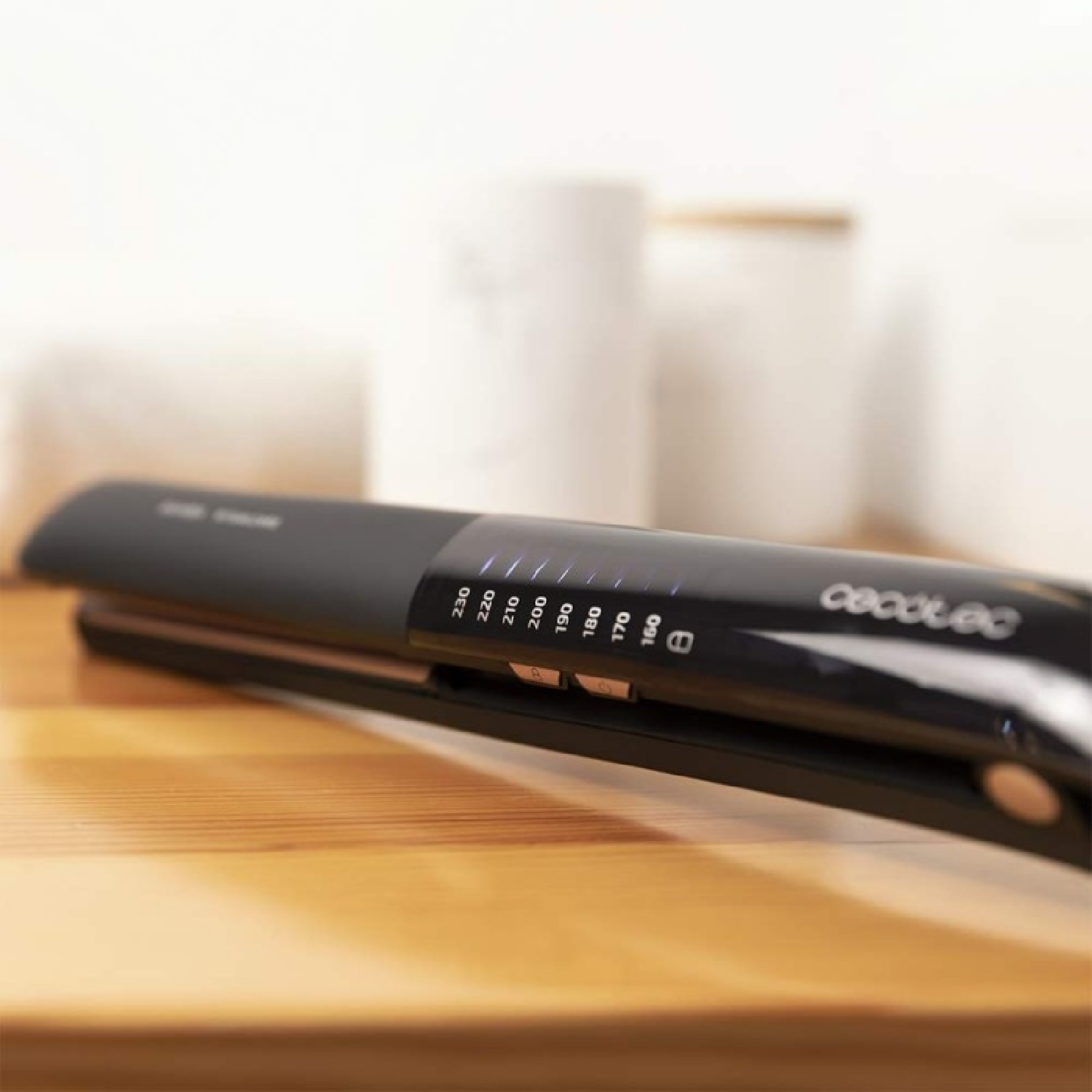Una piastra per capelli nera, con impostazioni di temperatura, è appoggiata su una superficie di legno. Due oggetti cilindrici bianchi sono sfocati sullo sfondo.