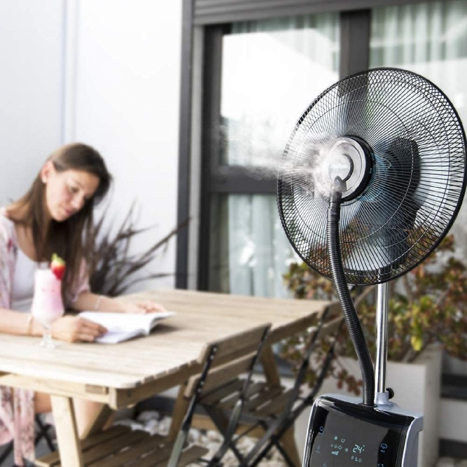 Kobieta czyta przy stole z napojem. Wiatrak mgiełkowy wieje. Rośliny i okno w tle.