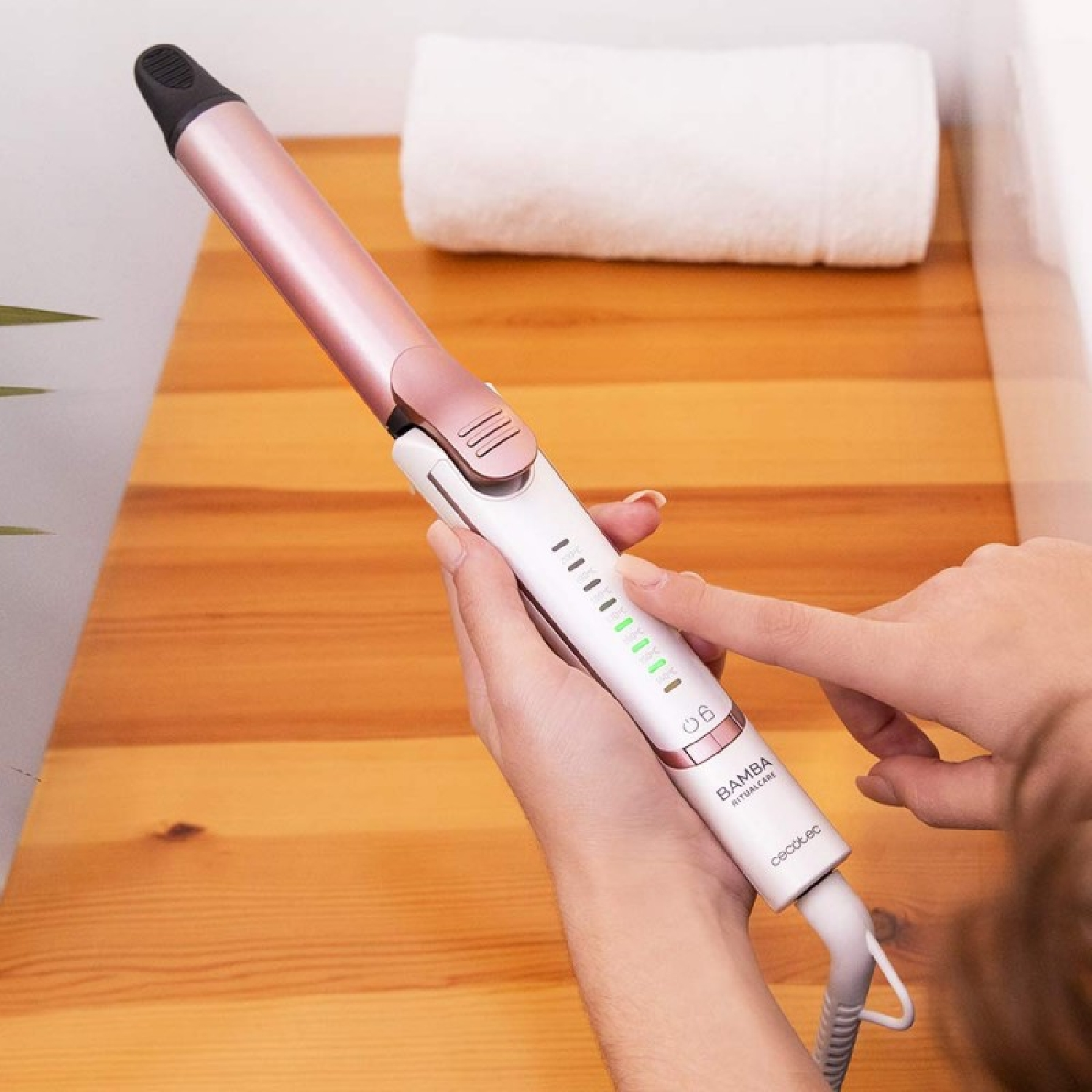 Osoba używa lokówki w kolorze różowo-białym na drewnianej powierzchni. Biały ręcznik w tle.