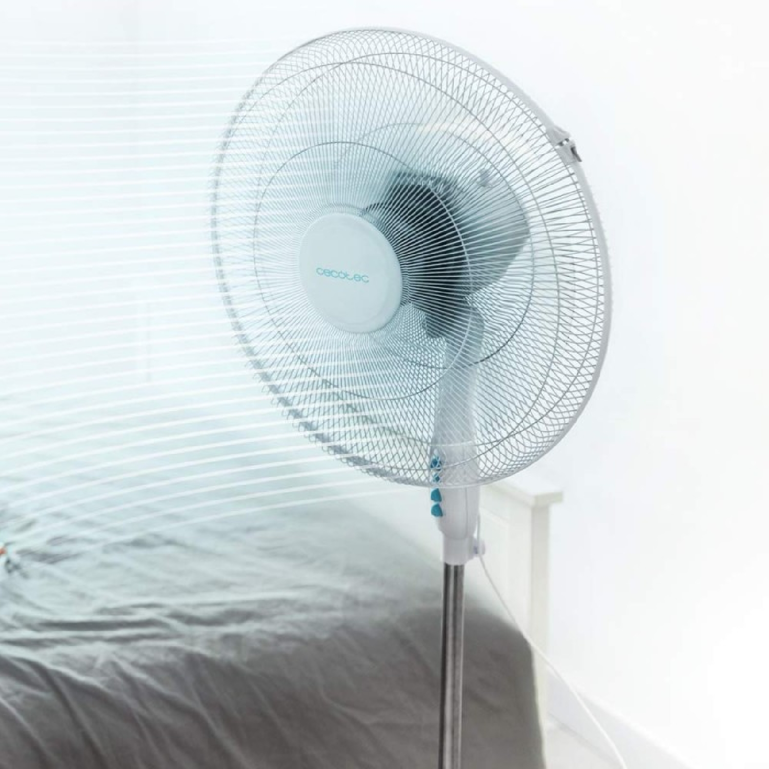 Biały i srebrny wentylator stojący znajduje się w pokoju. Wentylator znajduje się w pobliżu łóżka z szarą kołdrą. Tło to biała ściana.