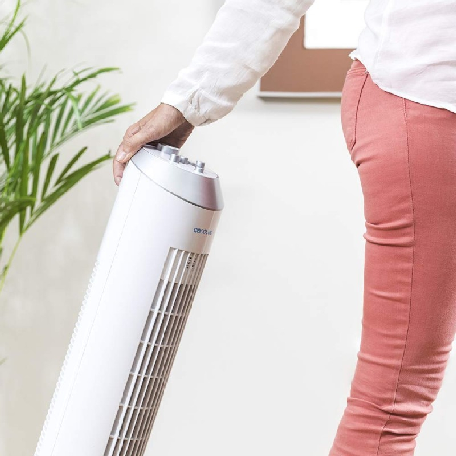 Osoba trzyma biały wentylator wieżowy, ubrana w białą koszulę i różowe spodnie. Zielona roślina w tle, brązowa rama obrazu.