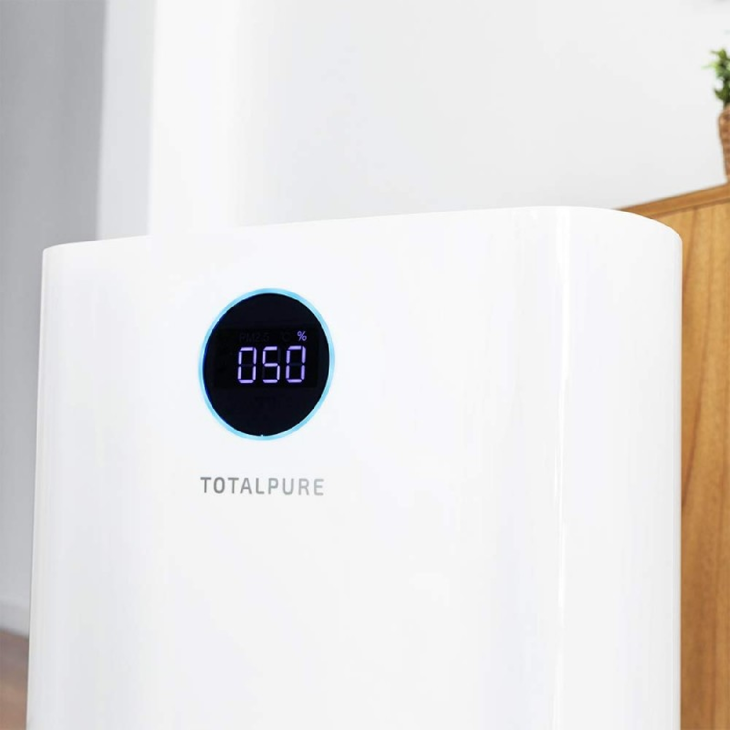 Un purificatore d'aria bianco mostra '050' in un cerchio blu, con 'TOTALPURE' sotto. Sullo sfondo una mensola in legno e una pianta.
