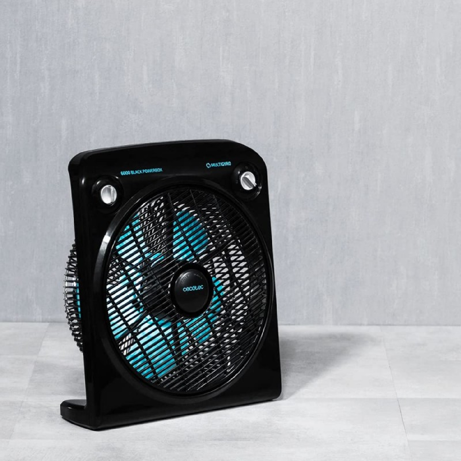 Czarny wentylator skrzynkowy z niebieskimi łopatkami stoi na szarej podłodze wyłożonej kafelkami. Wentylator jest lekko pochylony. Tłem jest szara ściana.