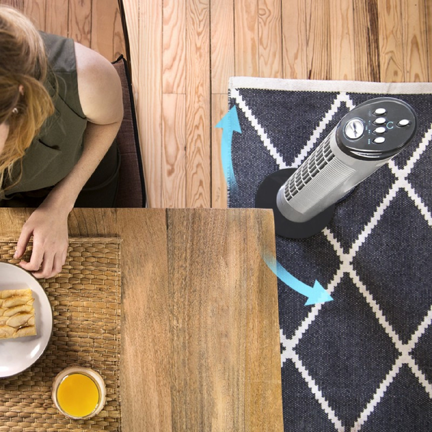 Kobieta siedzi przy drewnianym stole z jedzeniem i piciem. Wentylator stoi na dywanie. Strzałki wskazują przepływ powietrza.