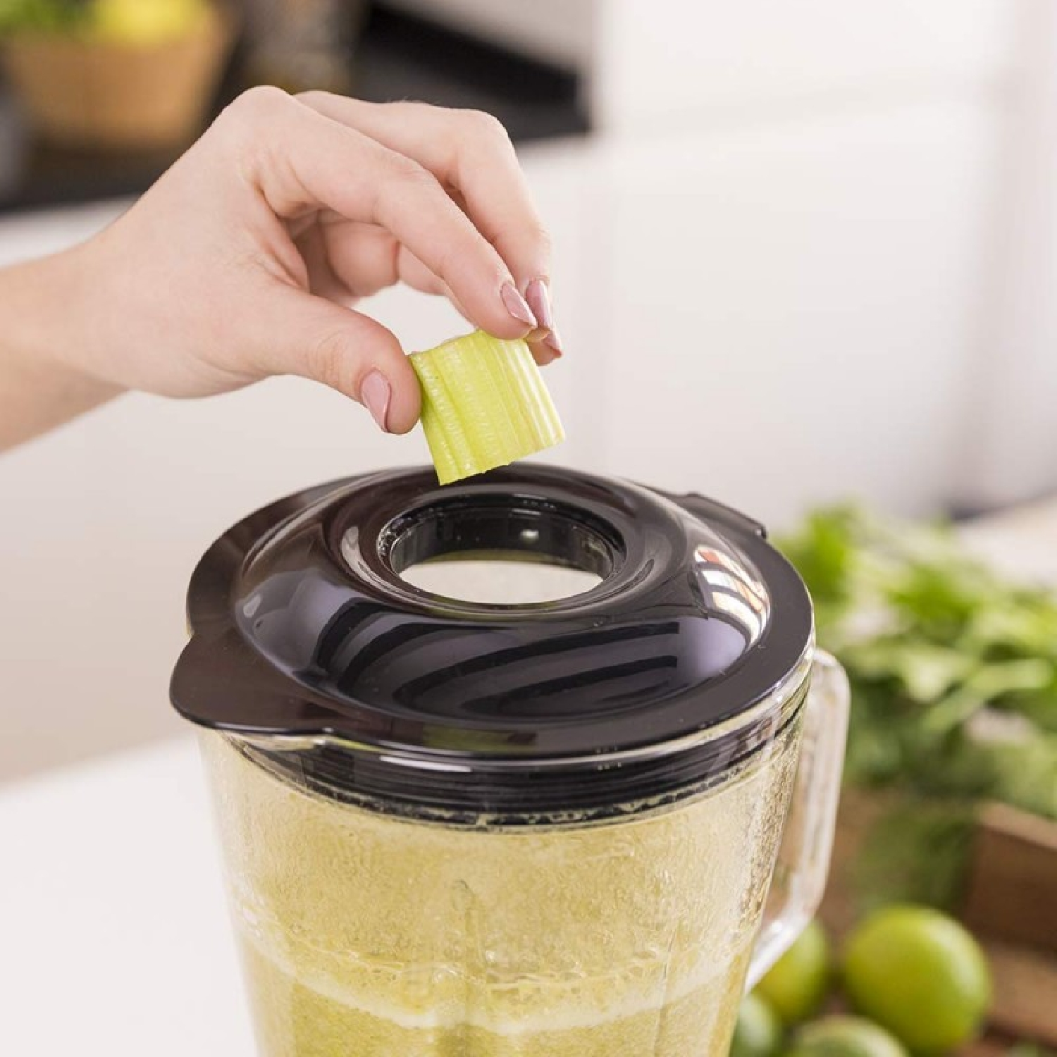 Una mano mette un pezzo di sedano in un frullatore. Il frullatore è pieno di liquido verde. Altri ingredienti e una scatola di legno sono sullo sfondo.