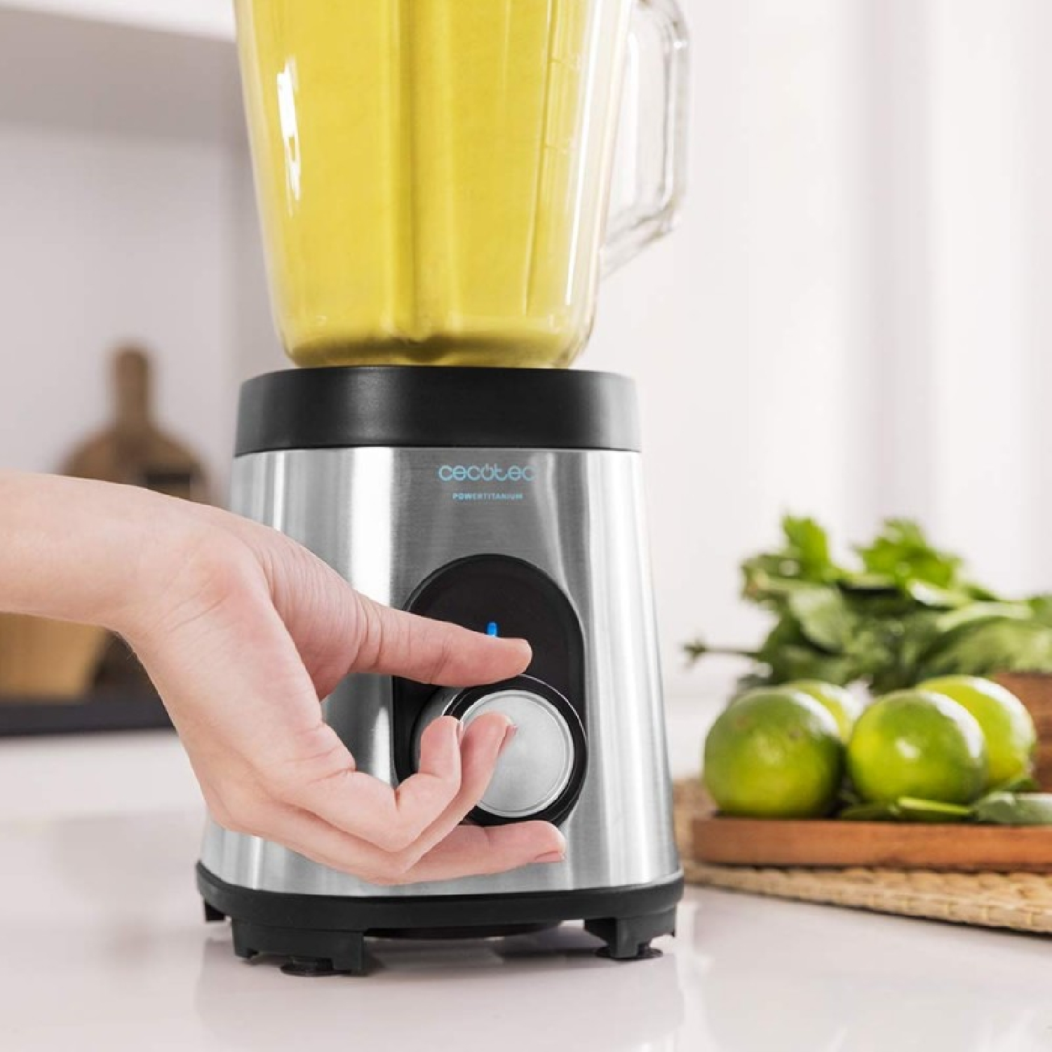 Una mano gira la manopola di un frullatore. Il frullatore ha una base argentata e un barattolo di vetro con liquido giallo. Lime ed erbe sono vicine.