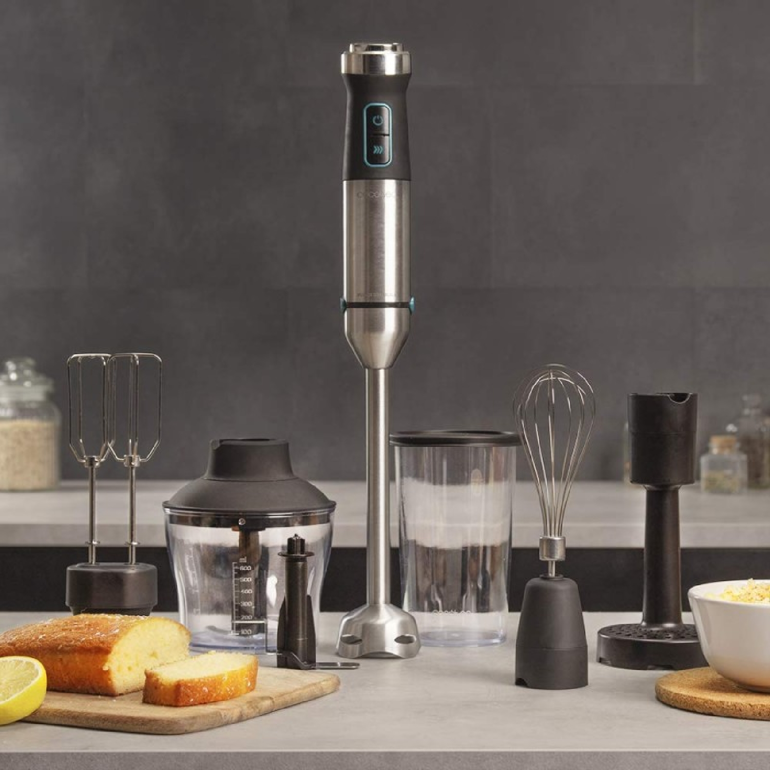 Una scena di cucina con un frullatore a immersione e accessori. Include frusta, sbattitore, tritatutto e misurino, con torta e limone.