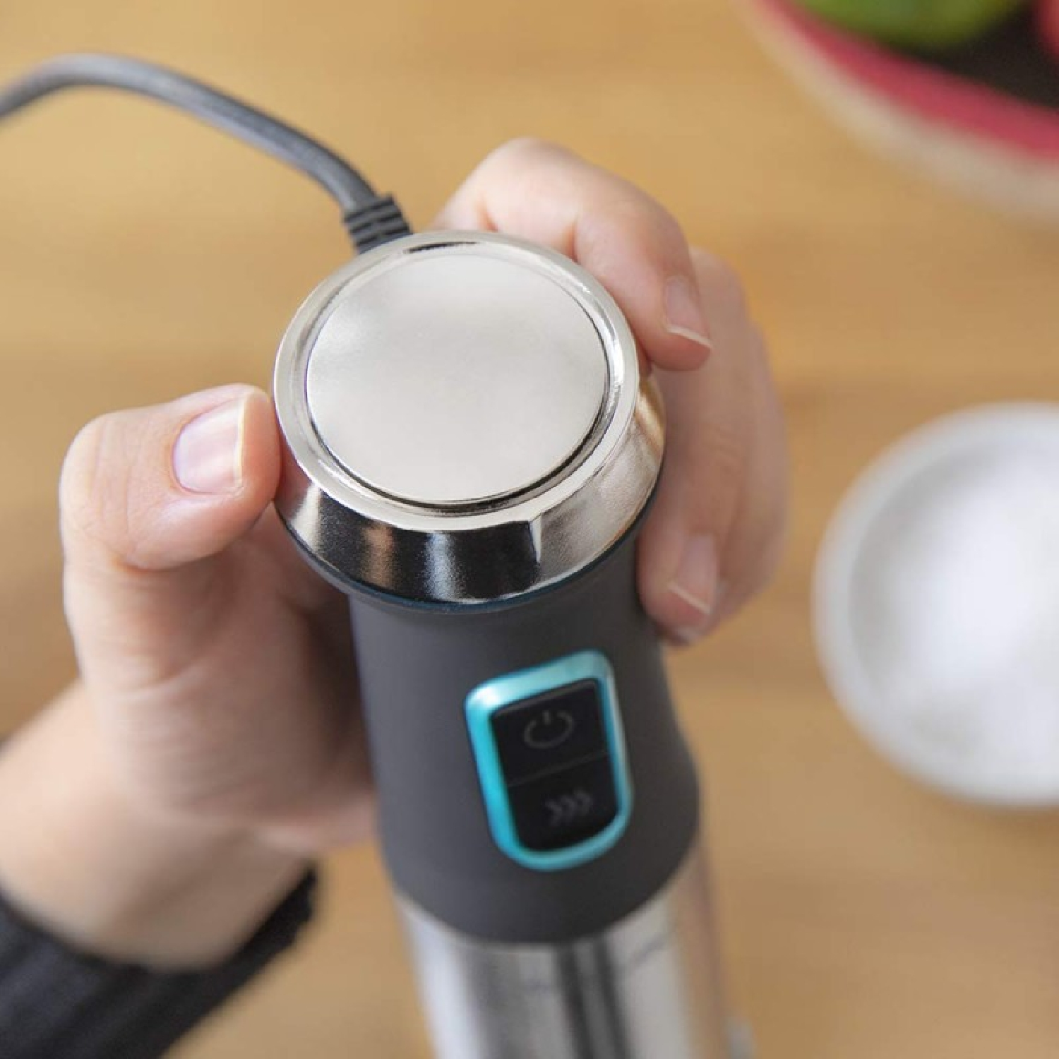 Dłoń trzymająca czarno-srebrny blender. Biała miska jest rozmyta w tle. Widoczna jest drewniana powierzchnia.