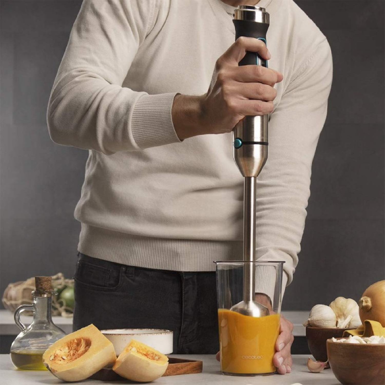 Een persoon gebruikt een staafmixer om oranje soep in een glas te mengen. Er liggen pompoenstukjes, knoflook en olie op tafel.