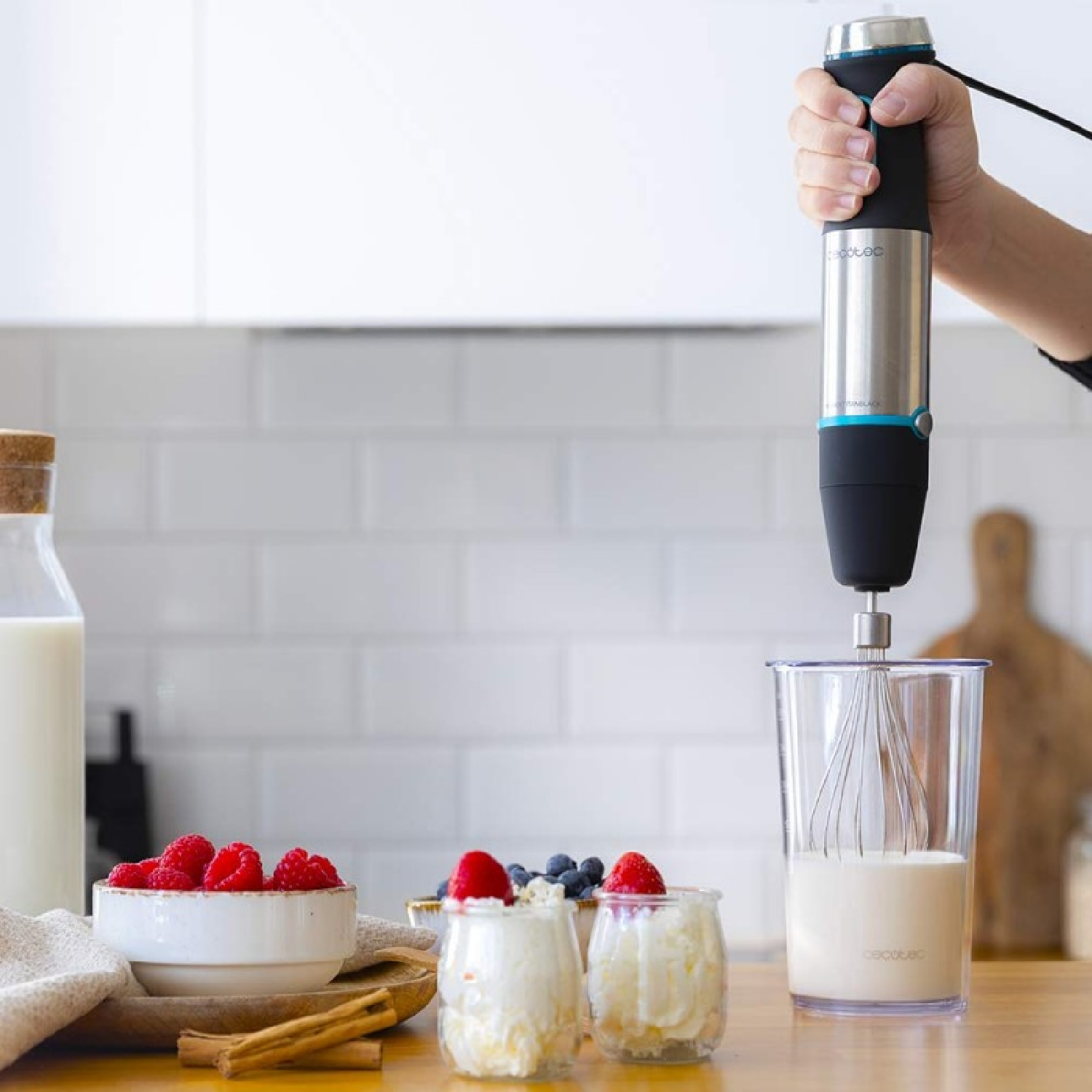 Eine Hand hält einen Mixer über einem Glas mit Schneebesen, Milch und Desserts mit Beeren auf einem Holztisch.