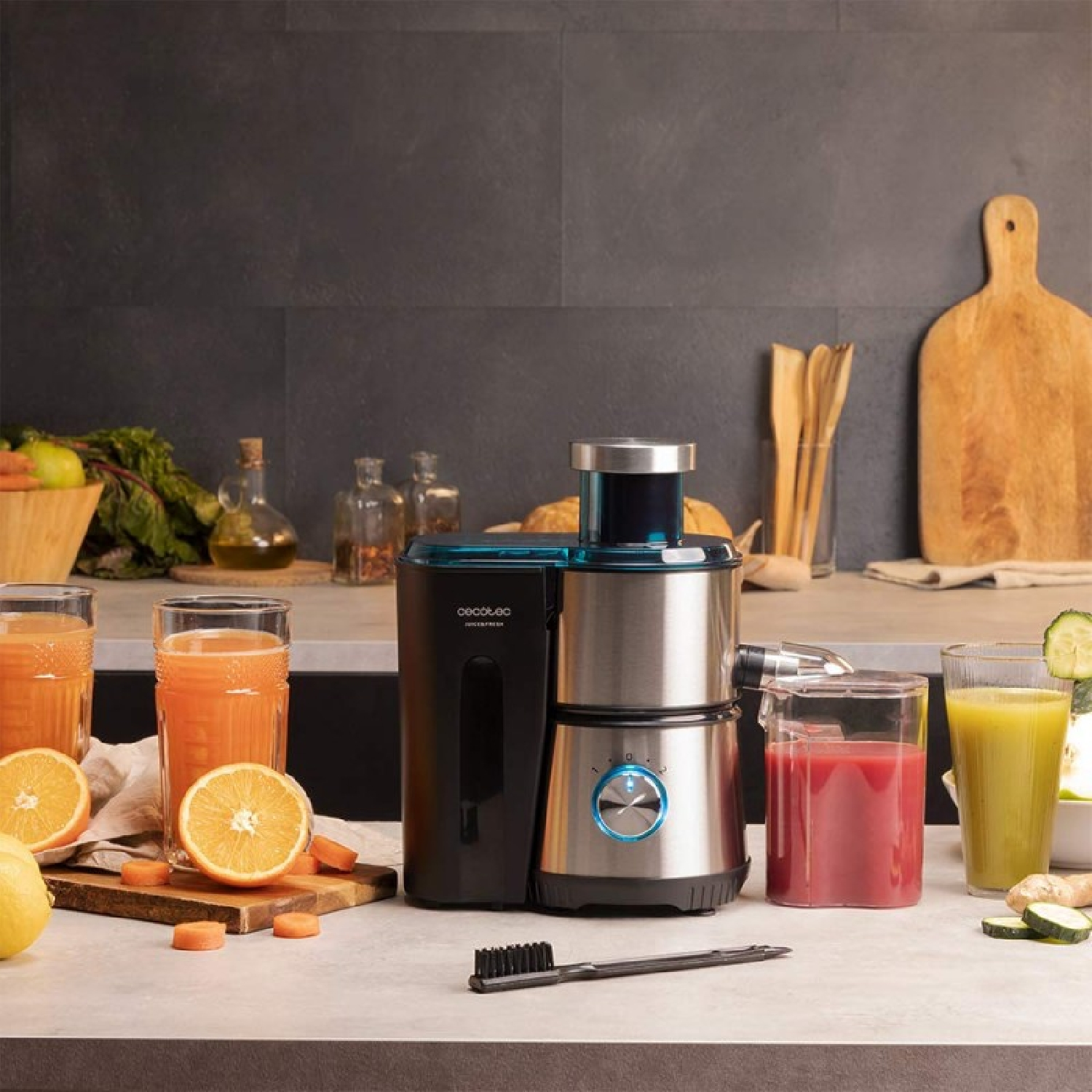 Una exprimidora con vasos de zumo, frutas y verduras. Se ven naranjas, zanahorias y rodajas de pepino.