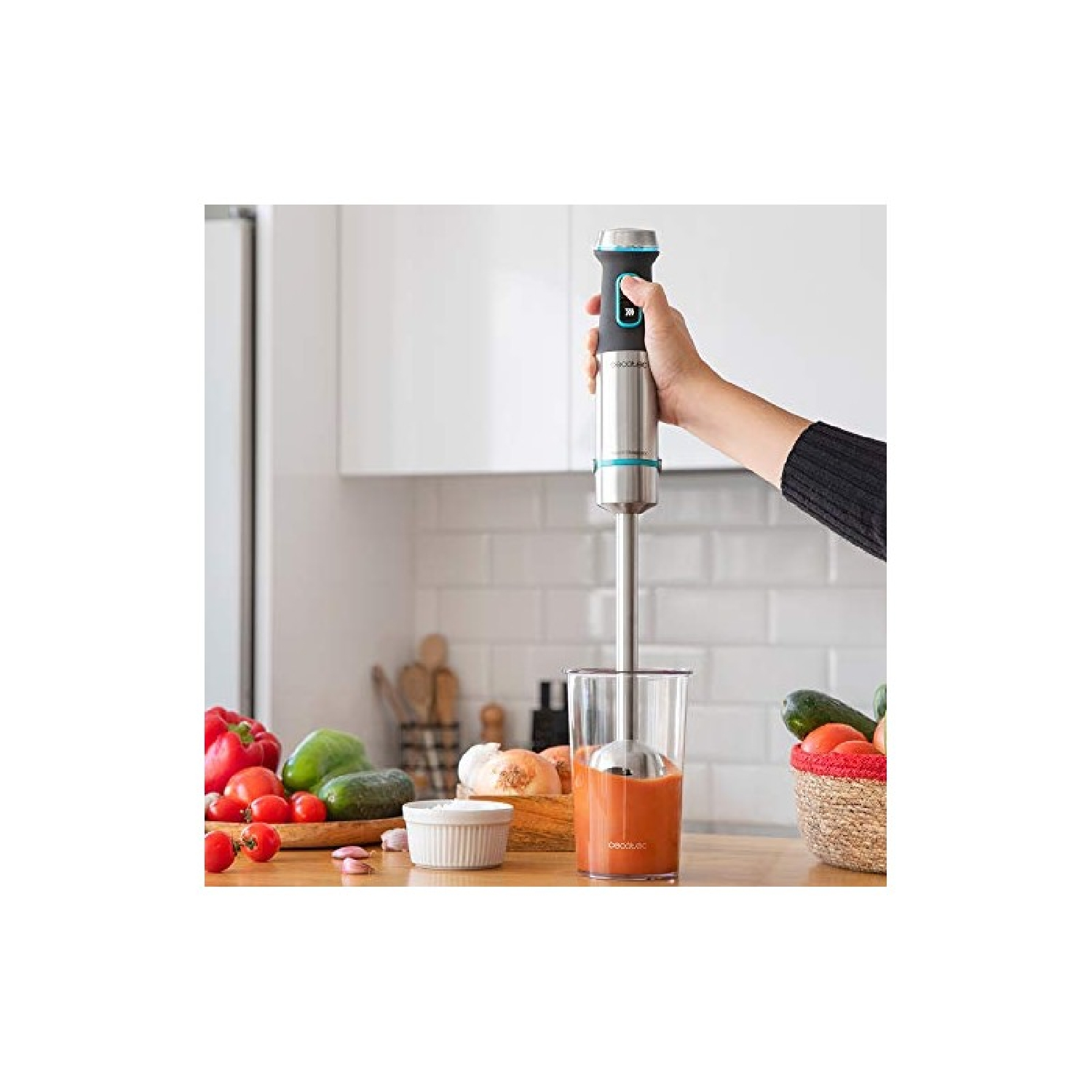 Een hand houdt een blender boven een glas oranje vloeistof. Groenten en een mand staan op tafel.