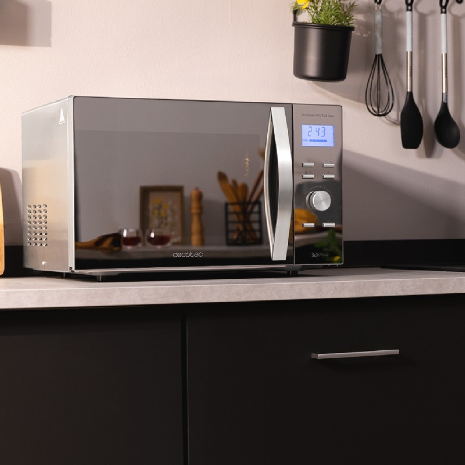 Un moderno horno microondas se encuentra sobre una encimera en una cocina. Utensilios cuelgan de la pared. Una pequeña planta encima del microondas.