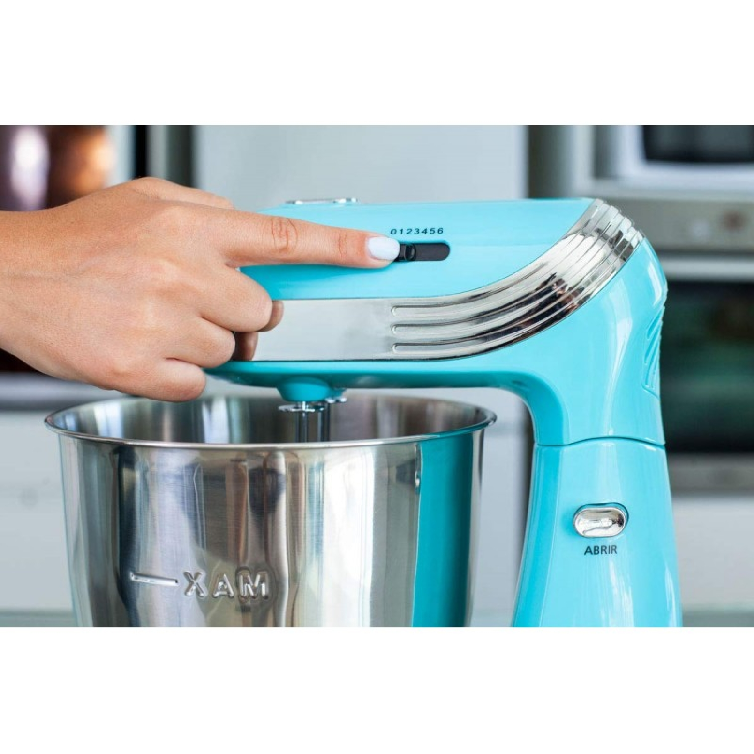 Una mano ajusta el dial de velocidad de una batidora de pie azul claro. Debajo hay un cuenco de acero inoxidable. El fondo es una cocina.