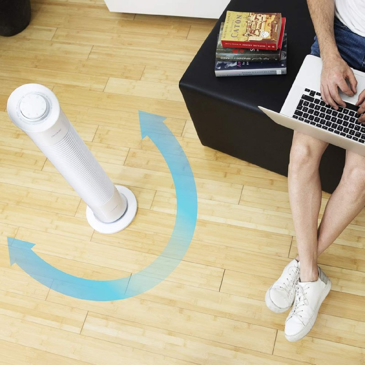 Osoba siedzi z laptopem w pobliżu białego wentylatora na drewnianej podłodze. Książki leżą na czarnej kostce. Niebieska strzałka wskazuje przepływ powietrza.