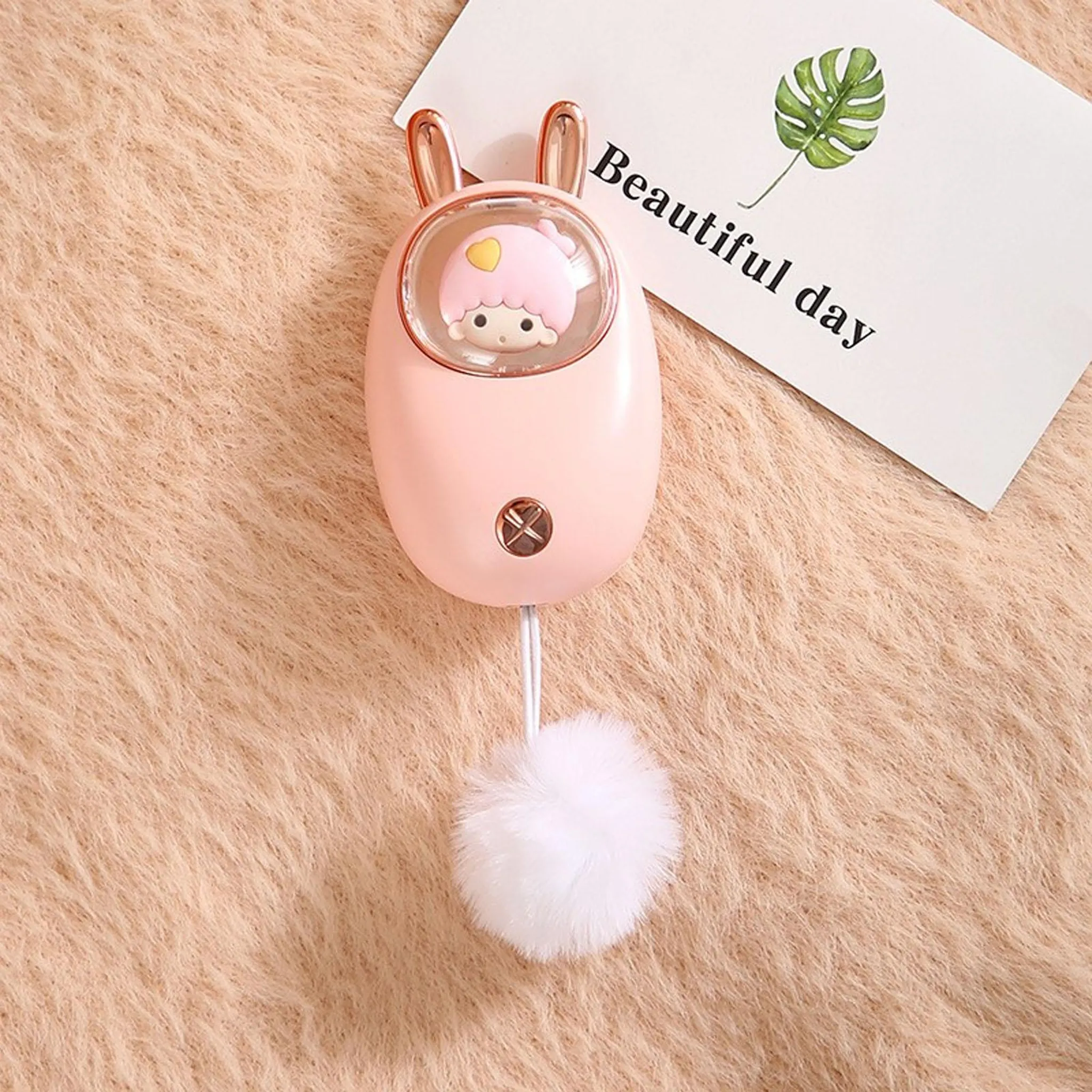Ein rosa, hasenförmiger Handwärmer mit einem flauschigen Schwanz und eine Karte mit einem Blatt und Text, alles auf einer flauschigen braunen Oberfläche.