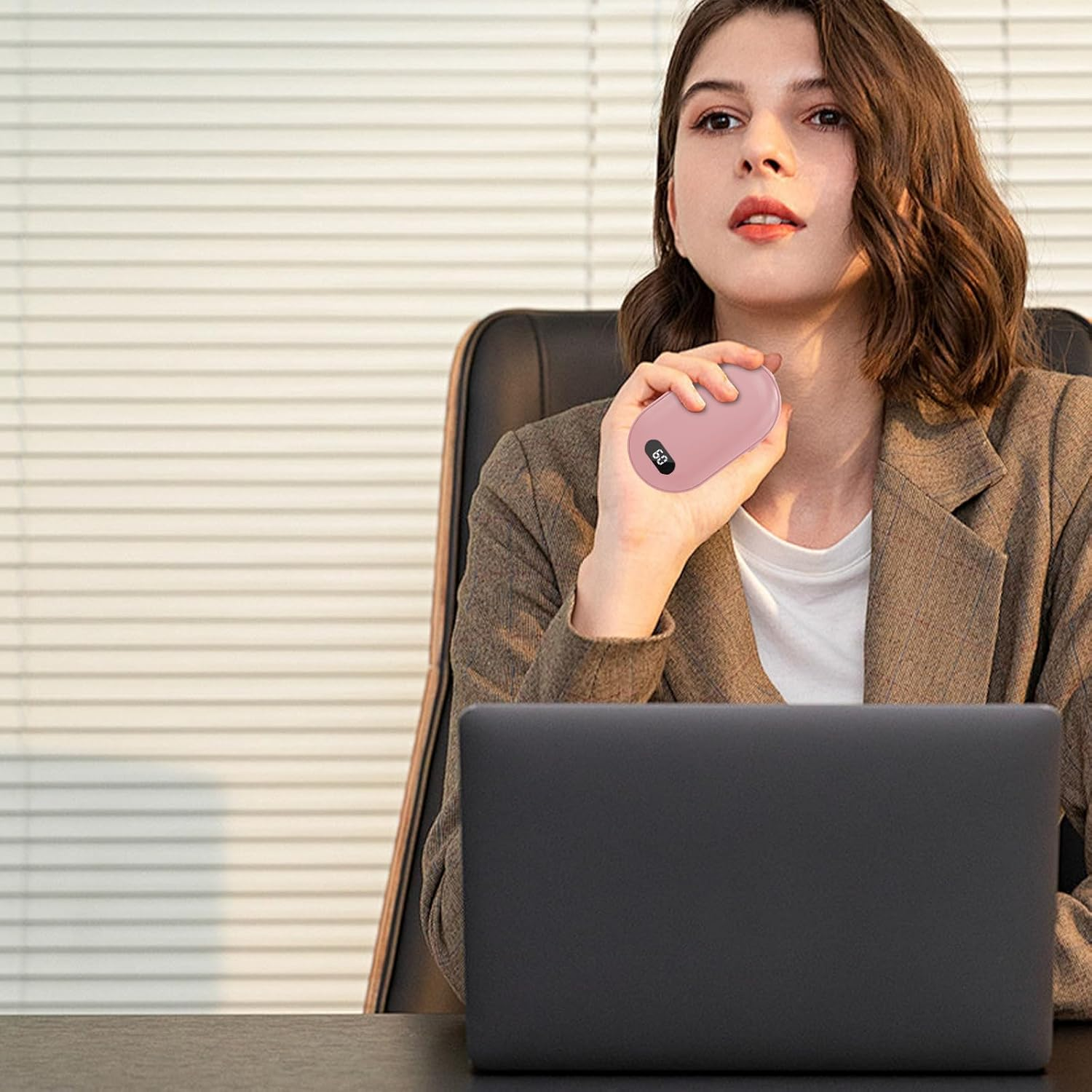 Eine Frau hält einen rosa Handwärmer und sitzt an einem Schreibtisch mit einem Laptop. Sie trägt einen Blazer und ein weißes Hemd.