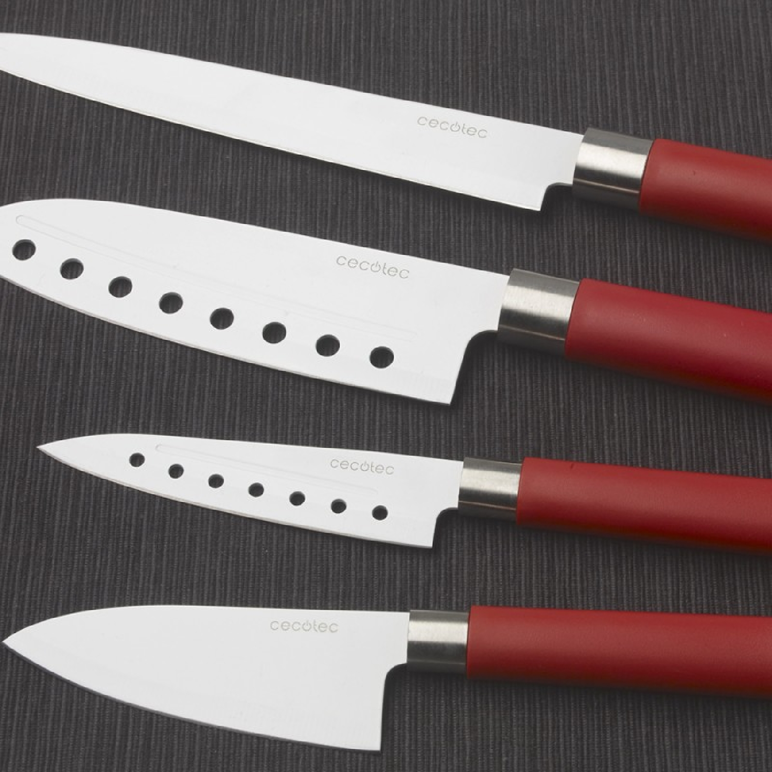 Cuatro cuchillos de cocina con hojas de acero inoxidable y mangos rojos, dispuestos diagonalmente sobre una superficie gris oscura texturizada.