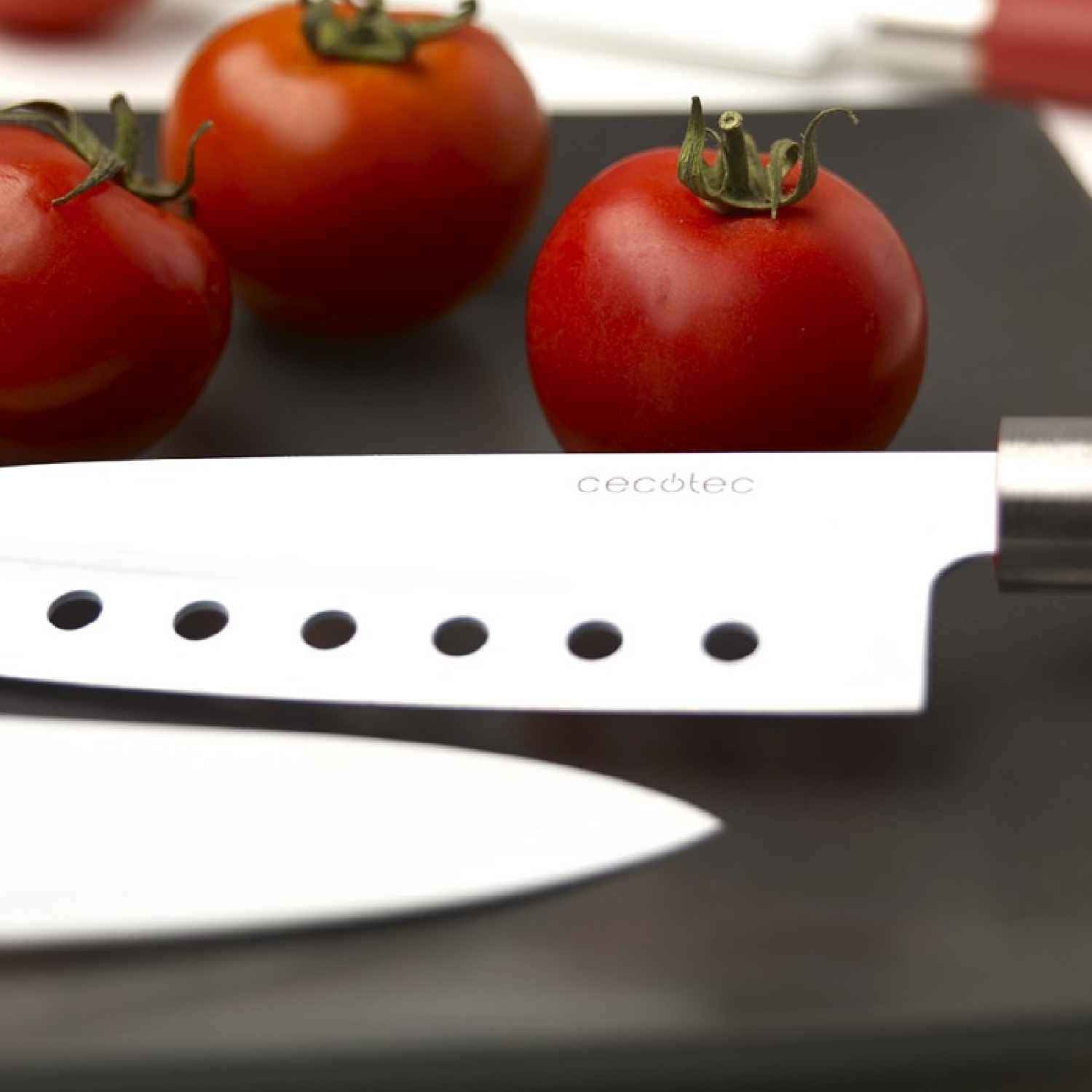 Tres tomates rojos con tallos verdes sobre una superficie negra. Un cuchillo plateado con agujeros al frente. Otro cuchillo debajo.
