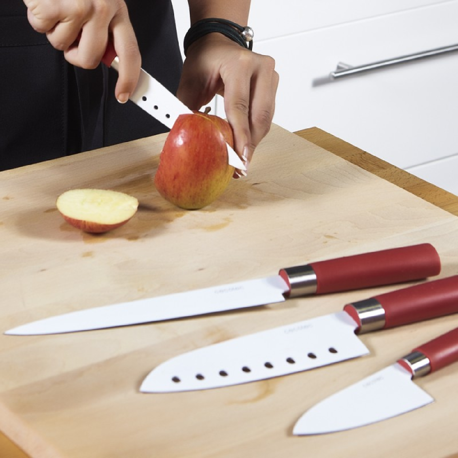 Una persona corta una manzana roja sobre una tabla de madera con un cuchillo blanco. Hay otros tres cuchillos con mangos rojos cerca.