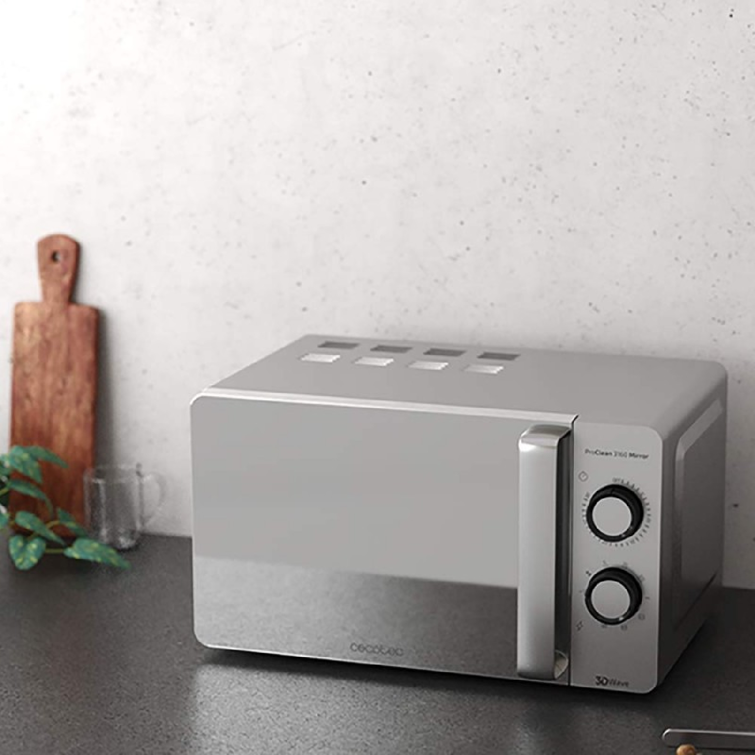 Srebrna kuchenka mikrofalowa na blacie. Drewniana deska do krojenia i szklanka po lewej stronie. Ściana jest biała.