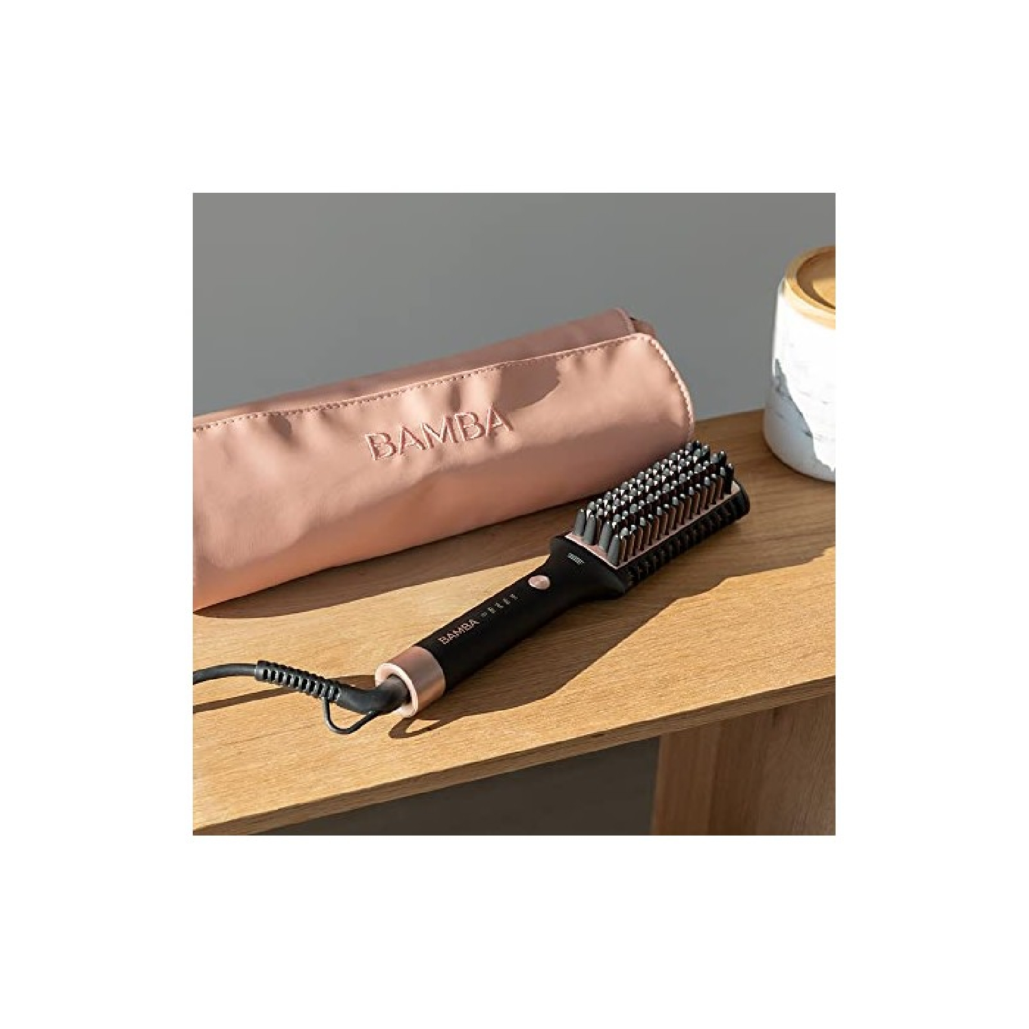 Een zwarte en roségouden haarstijlborstel, een roze etui met "BAMBA" erop en een witte pot met een houten deksel op een houten oppervlak.