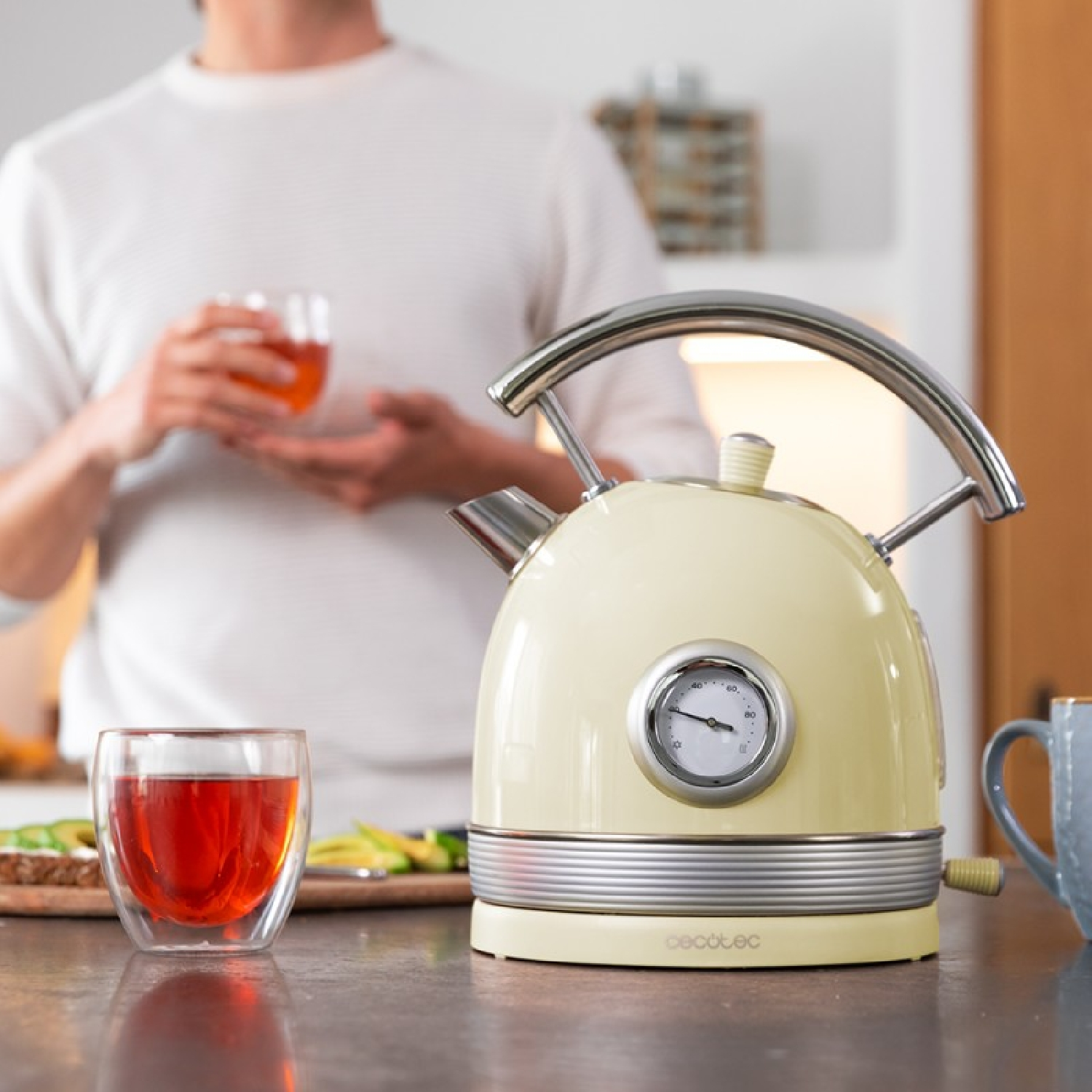 Una tetera crema con un indicador de temperatura está en una encimera. Una persona sostiene un vaso de té. Un vaso de té está en primer plano.