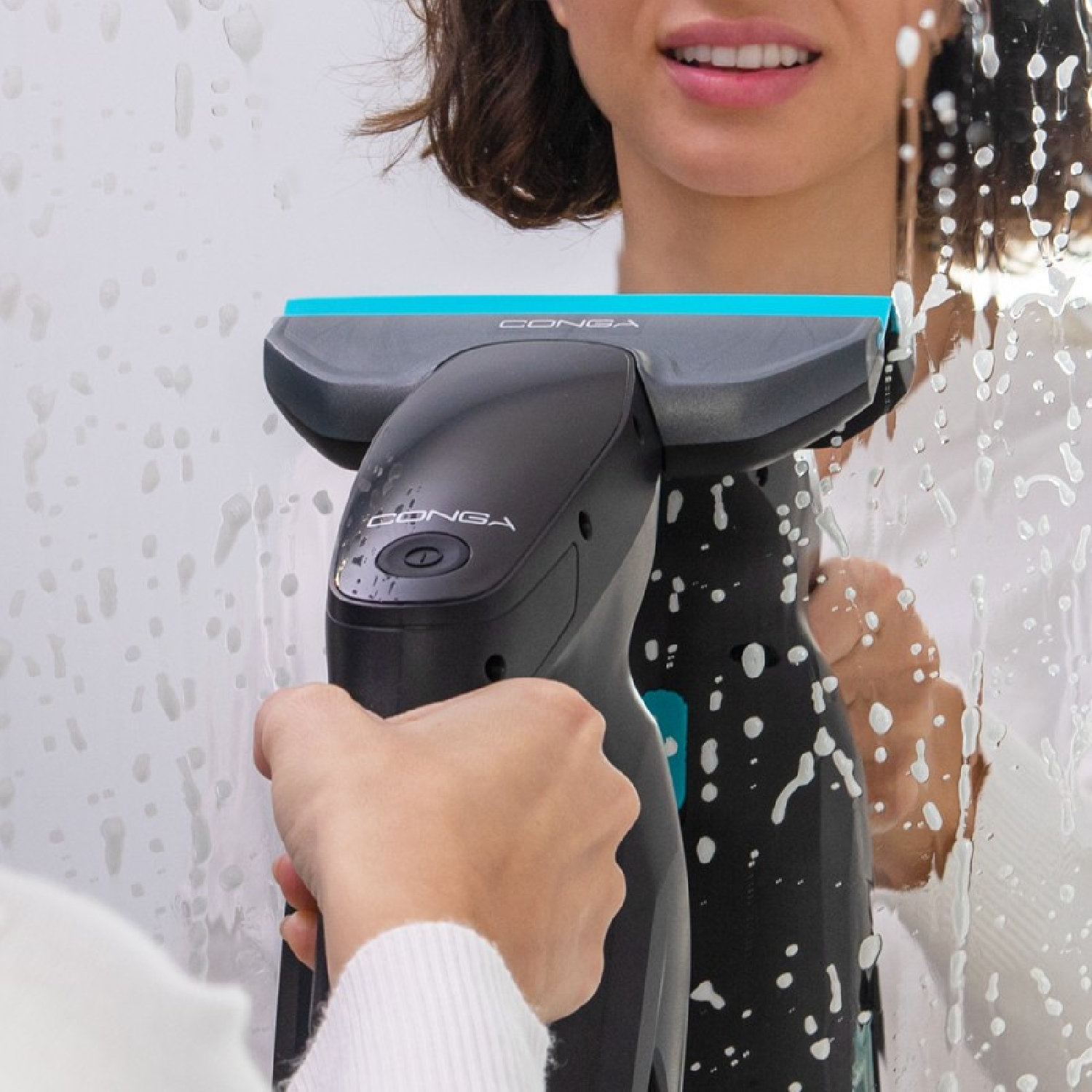 Kobieta używa czarnego i niebieskiego urządzenia do czyszczenia okien. Na urządzeniu jest napis 'CONGA'. Kobieta uśmiecha się, czyszcząc mydlane okno.