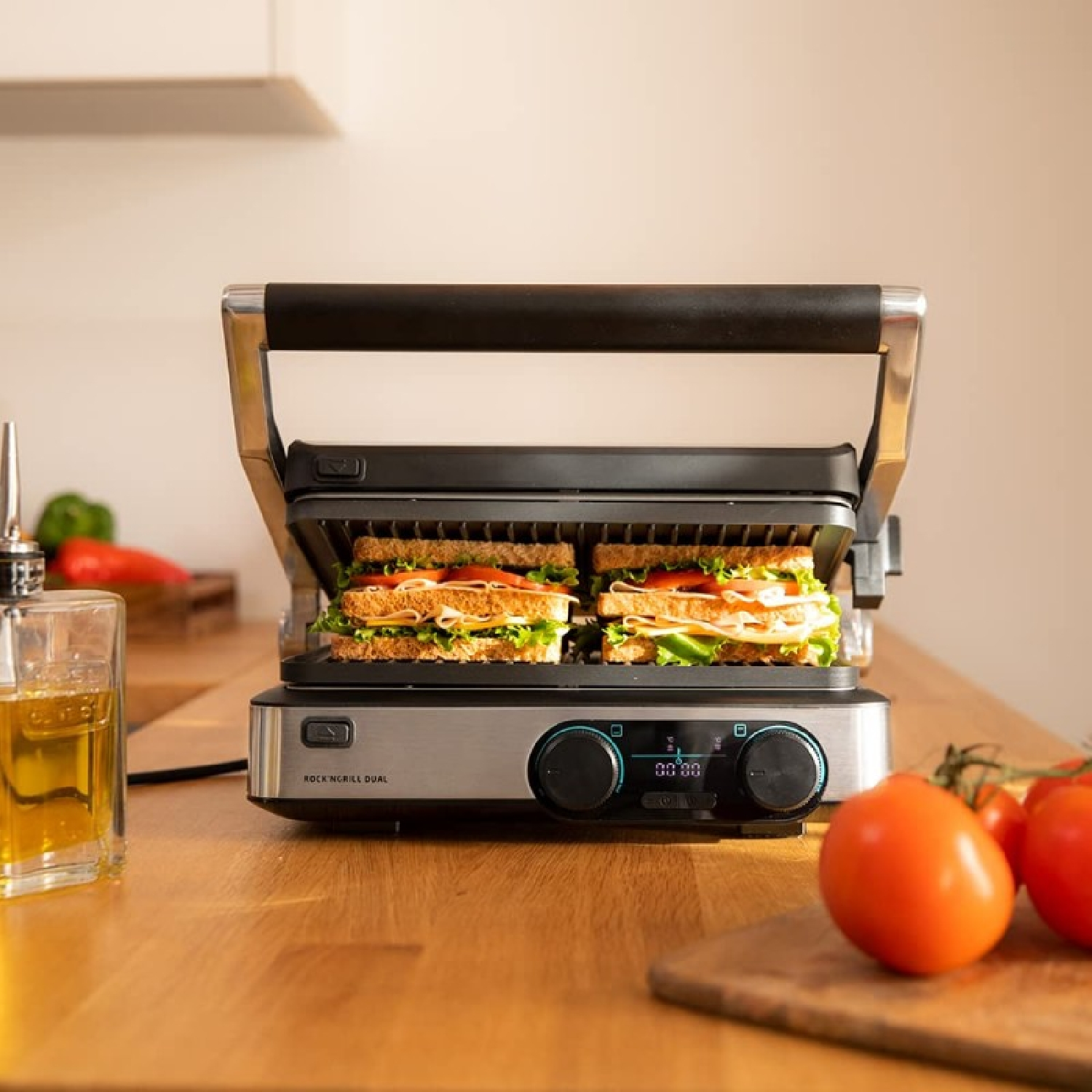 Una piastra per sandwich con due sandwich, pomodori, olio d'oliva e verdure su un bancone di legno in cucina.