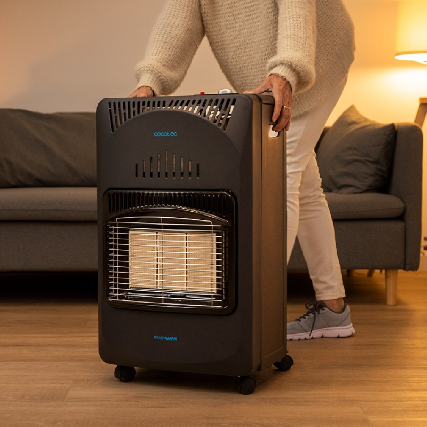 Eine Person hält einen schwarzen Gasheizer mit Heizelement, Rädern und dem Ready Warm-Logo. Im Hintergrund ist ein Sofa zu sehen.