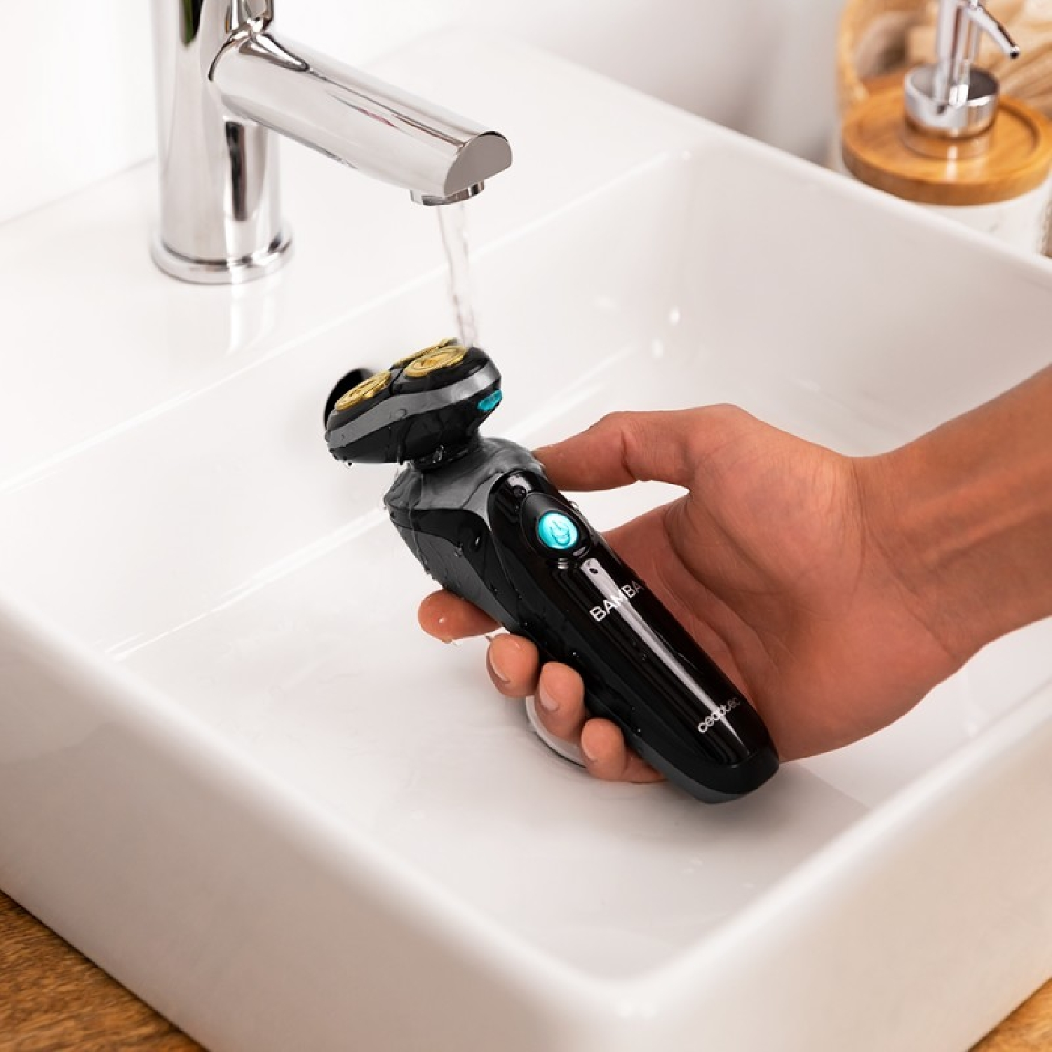Eine Hand hält einen schwarz-grauen Rasierer unter einem laufenden Wasserhahn in einem weißen Waschbecken. Der Rasierer wird gereinigt.