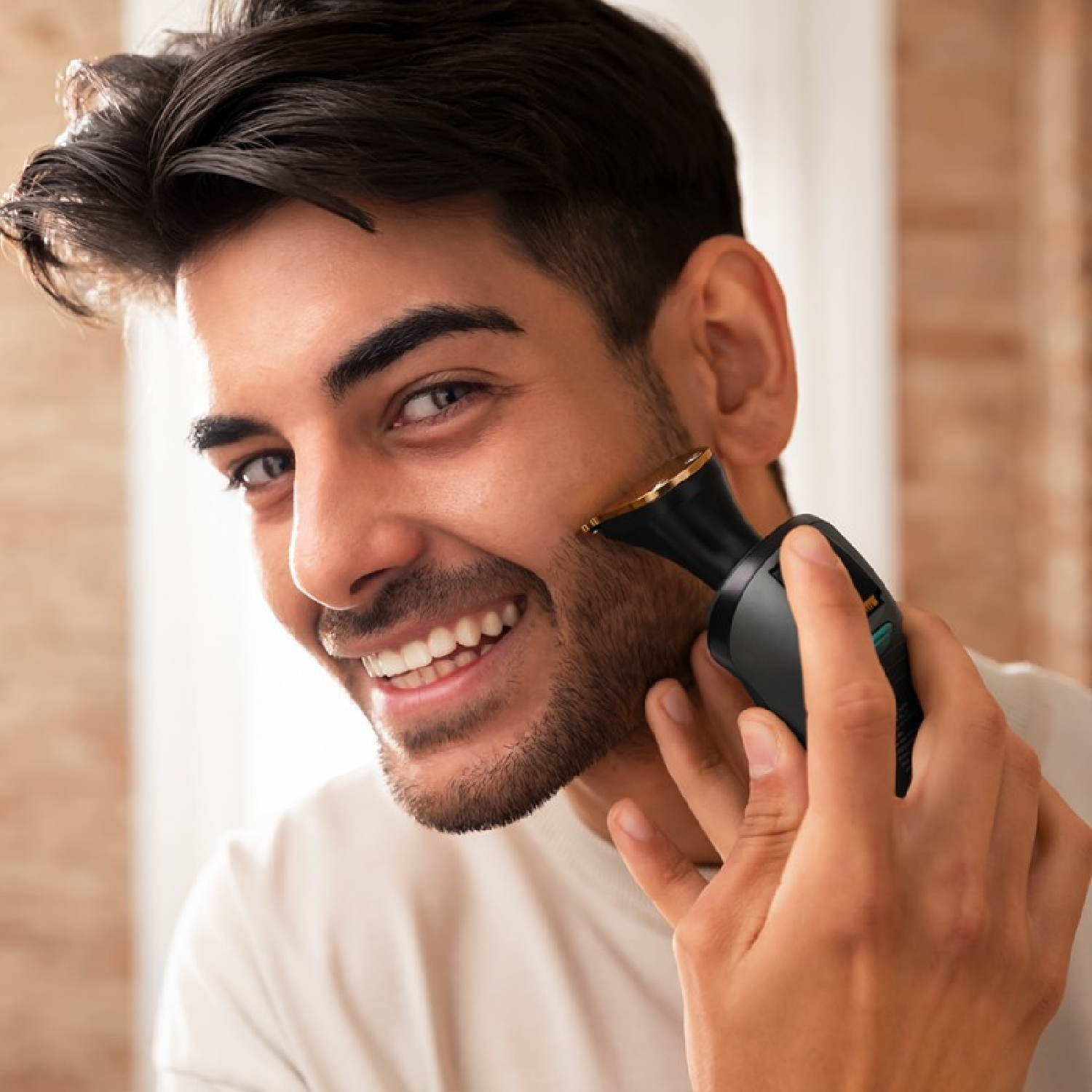 Ein lächelnder Mann rasiert seinen Bart mit einem schwarzen Trimmer. Er trägt ein weißes Hemd. Der Hintergrund ist unscharf.