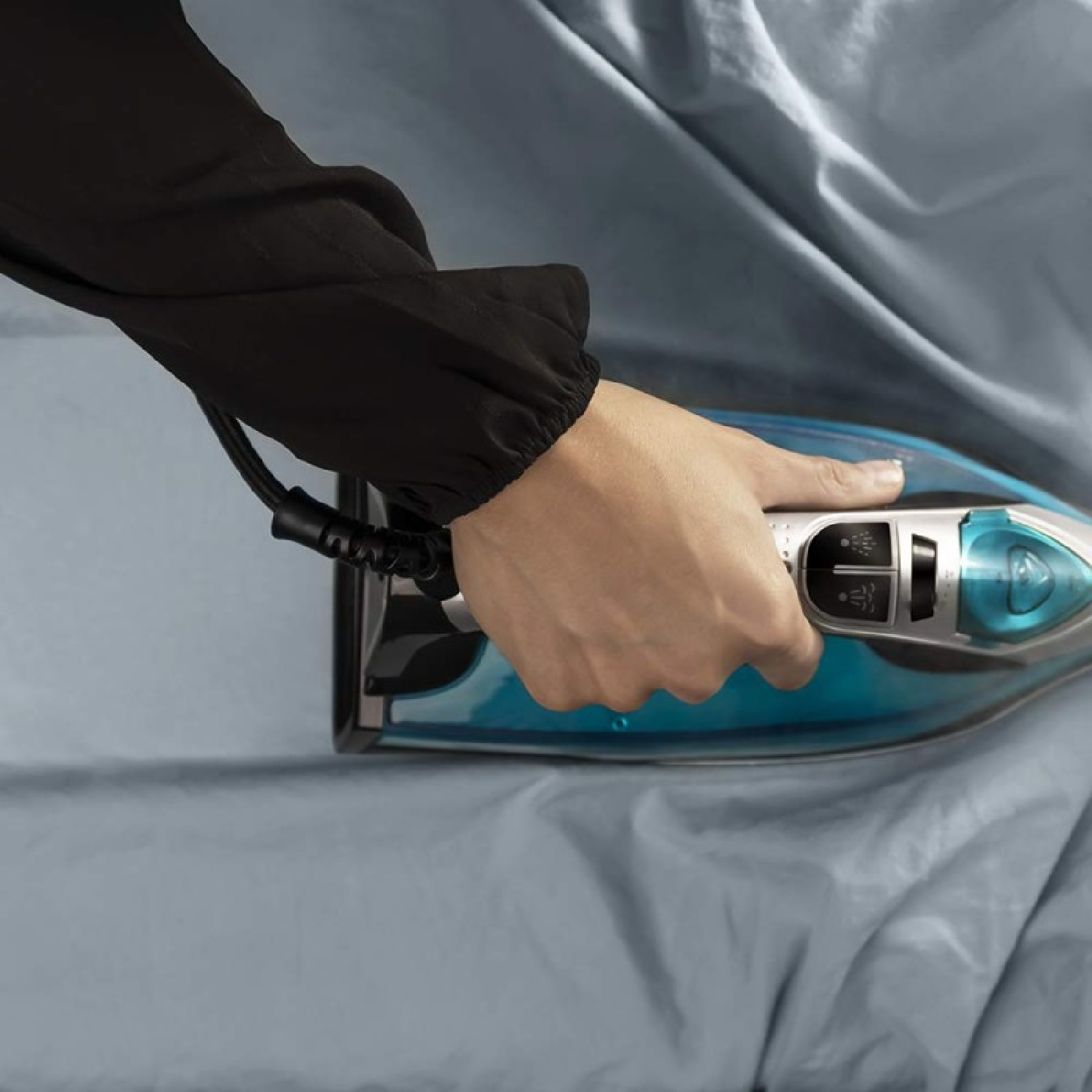 Eine Hand bügelt blauen Stoff mit einem blau-silbernen Bügeleisen. Die Person trägt einen schwarzen Ärmel. Der Stoff hat Falten.