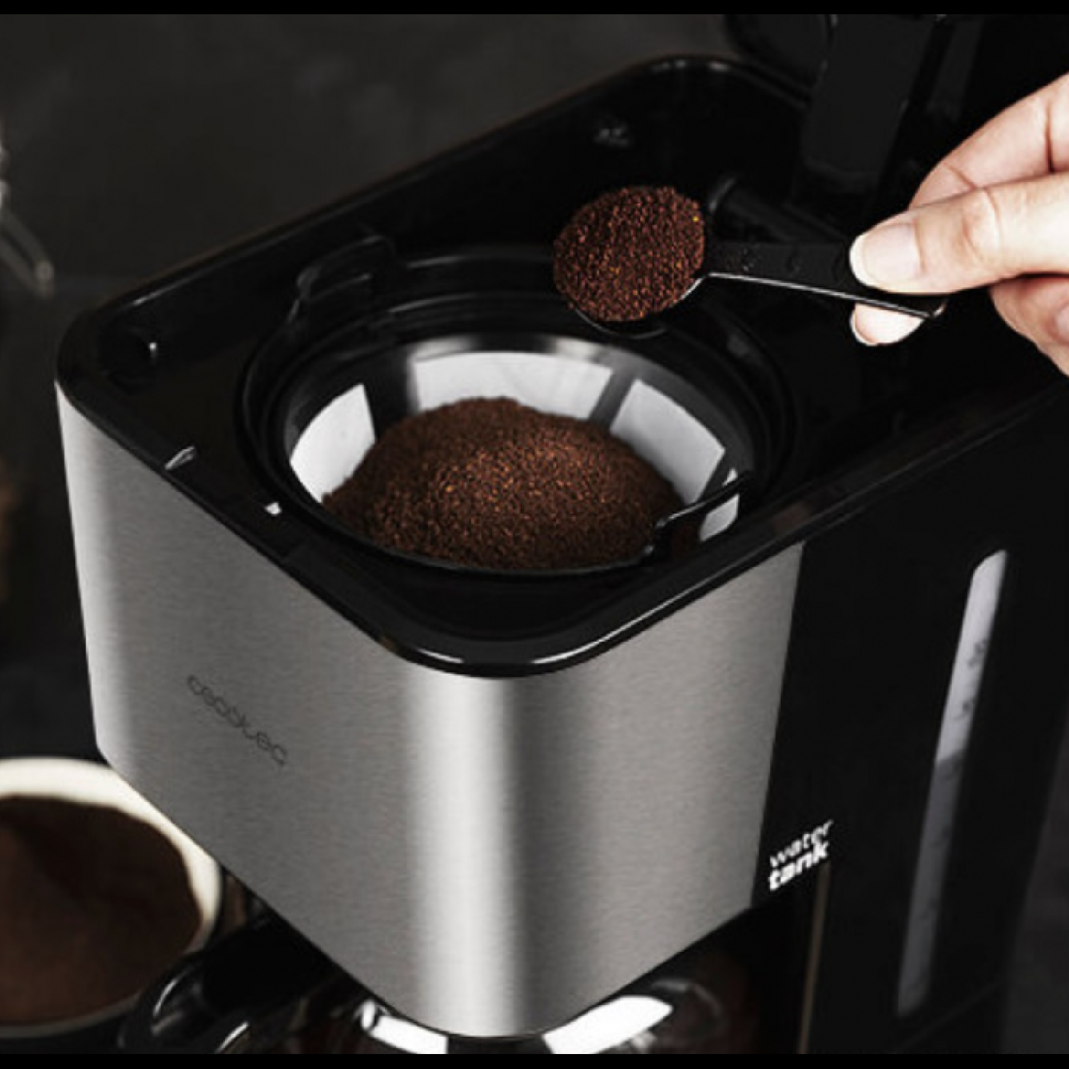 Ręka wsypuje zmieloną kawę do filtra ekspresu. Ekspres jest czarny i srebrny.