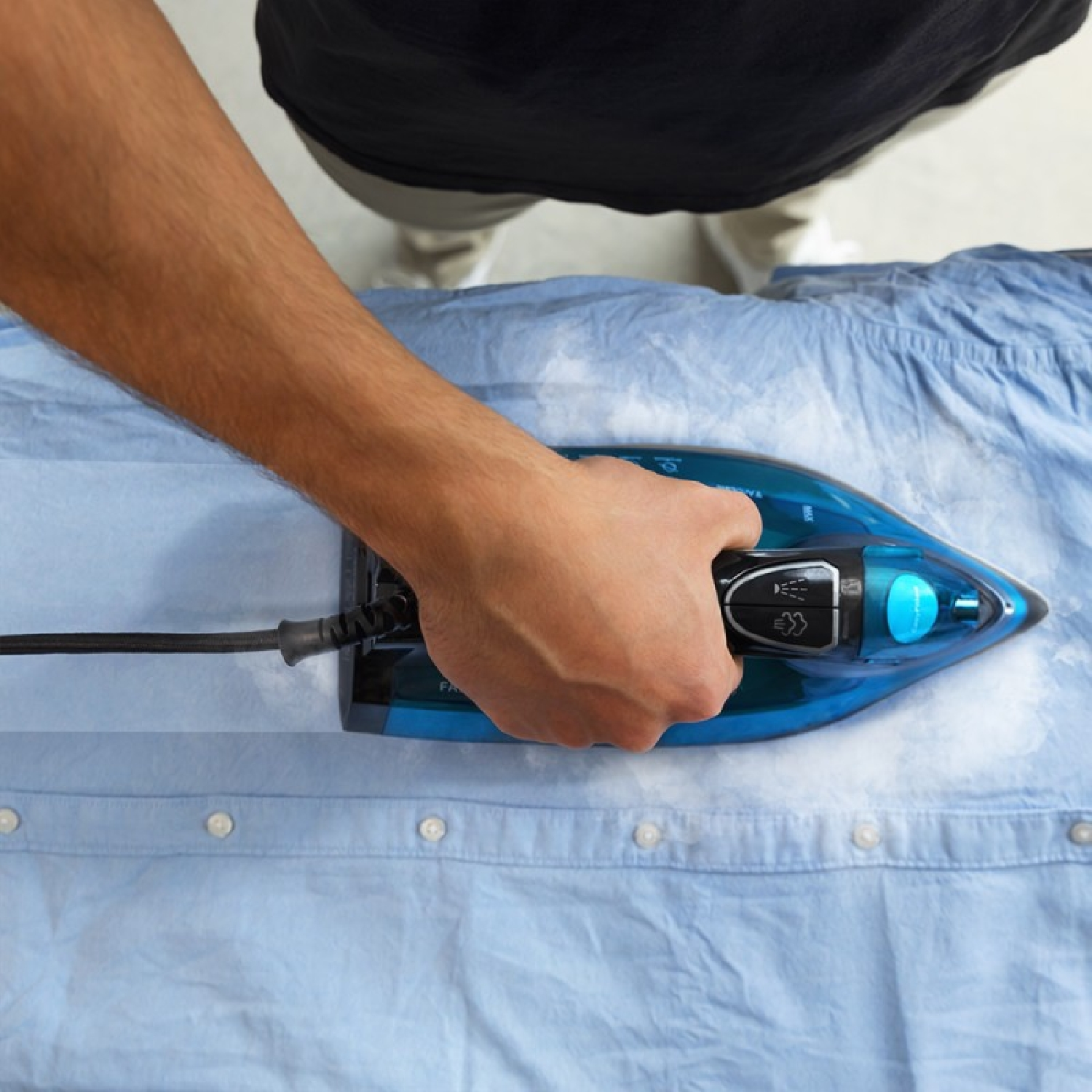 Osoba prasuje niebieską koszulę niebieskim żelazkiem. Żelazko styka się z koszulą, widoczna jest ręka osoby.