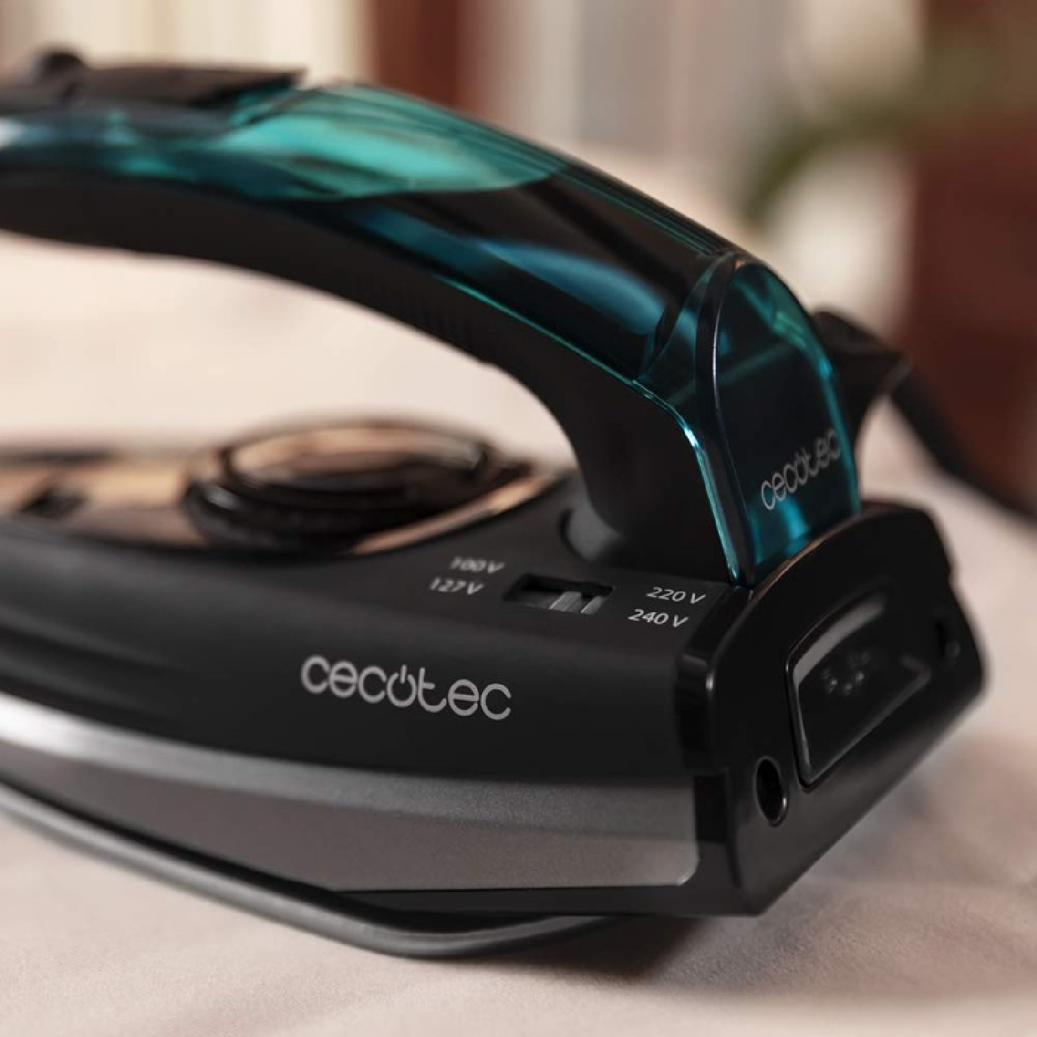 Zbliżenie czarnego i turkusowego żelazka z marką 'cecotec'. Żelazko spoczywa na białej powierzchni, rozmyte tło.