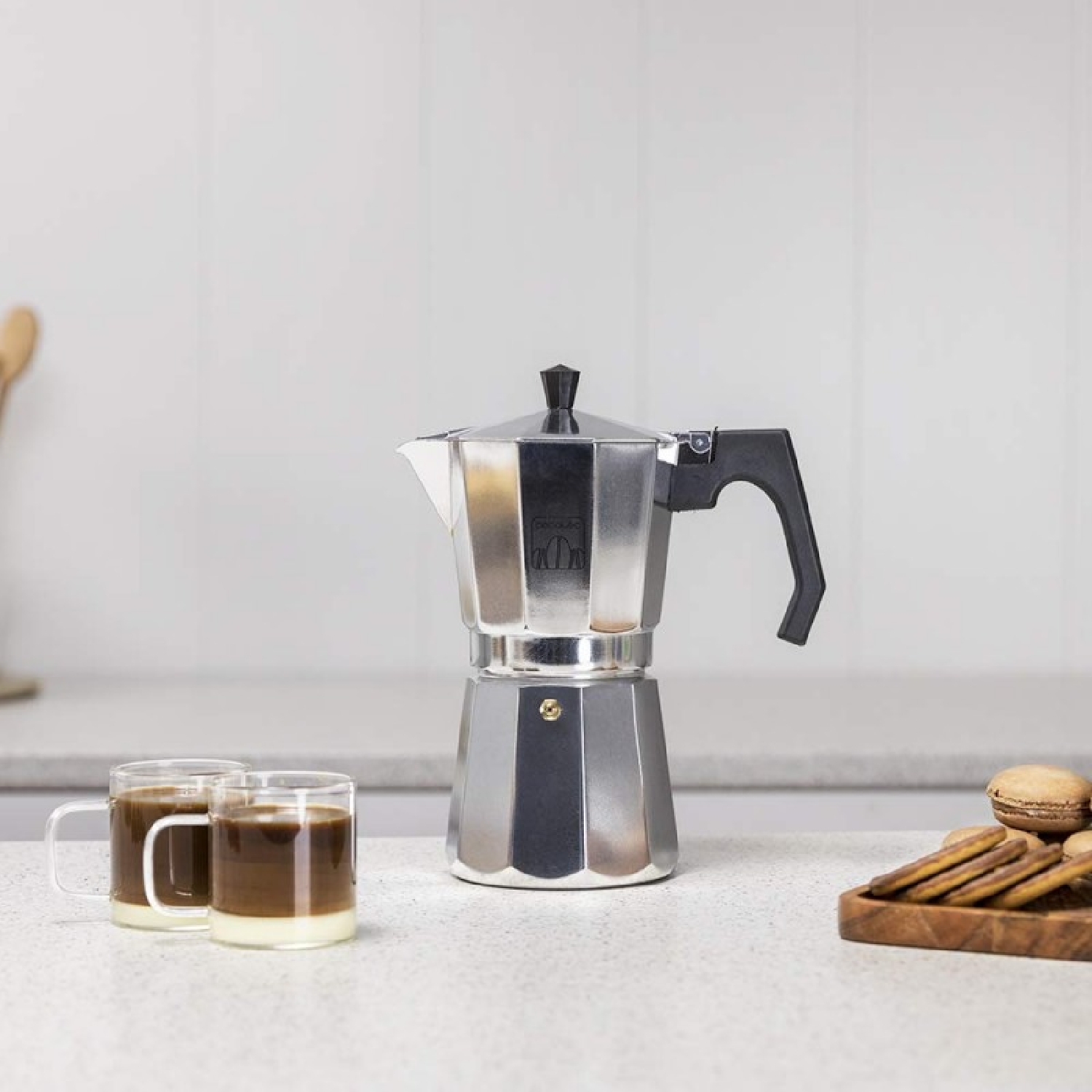 Ein Metall-Kaffeebereiter steht neben zwei Gläsern Kaffee mit Milch. Kekse und Makronen sind auf einem Holztablett.